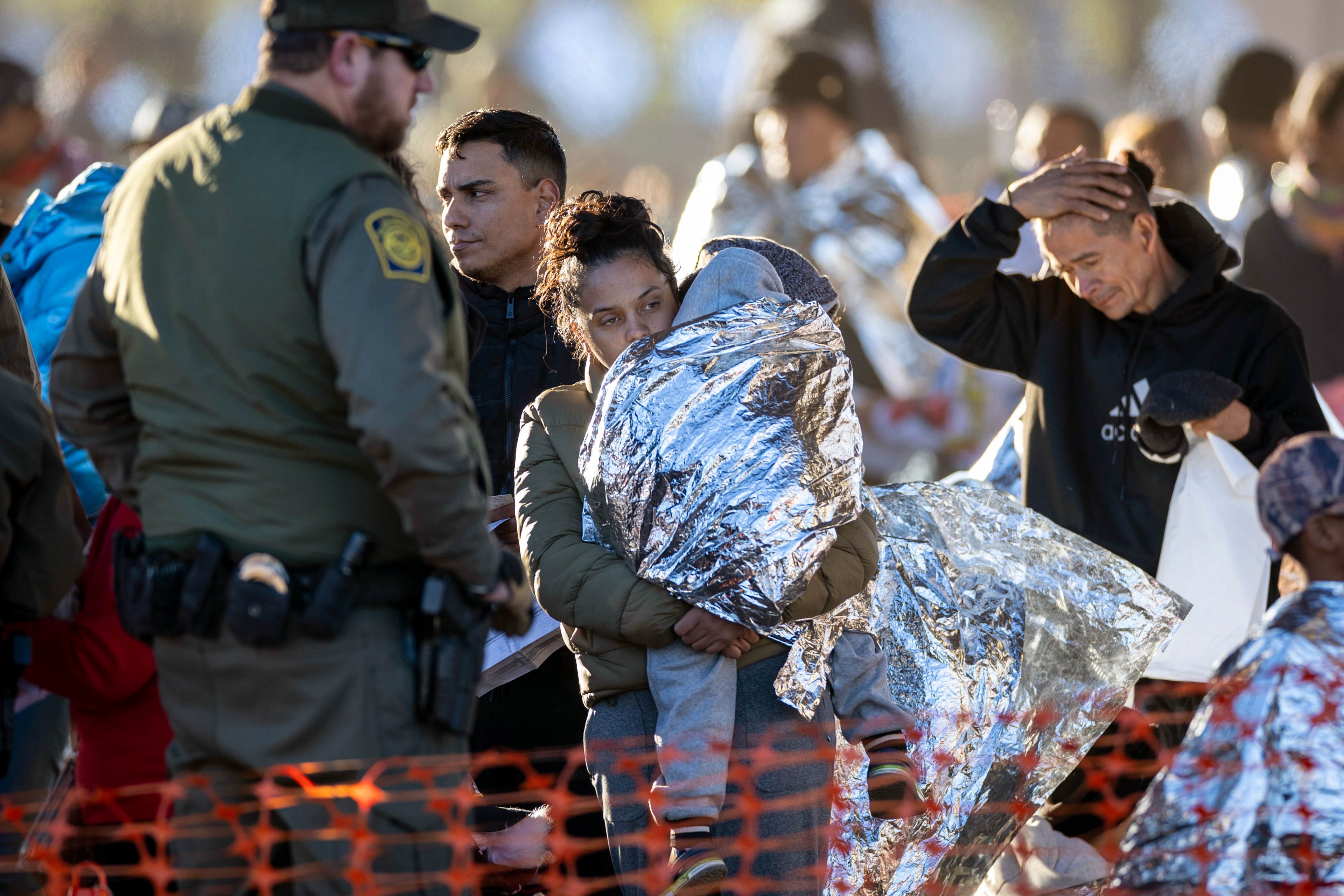 A woman holds a child wrapped in a reflective blanket, overseen by a border patrol officer. A man looks concerned.