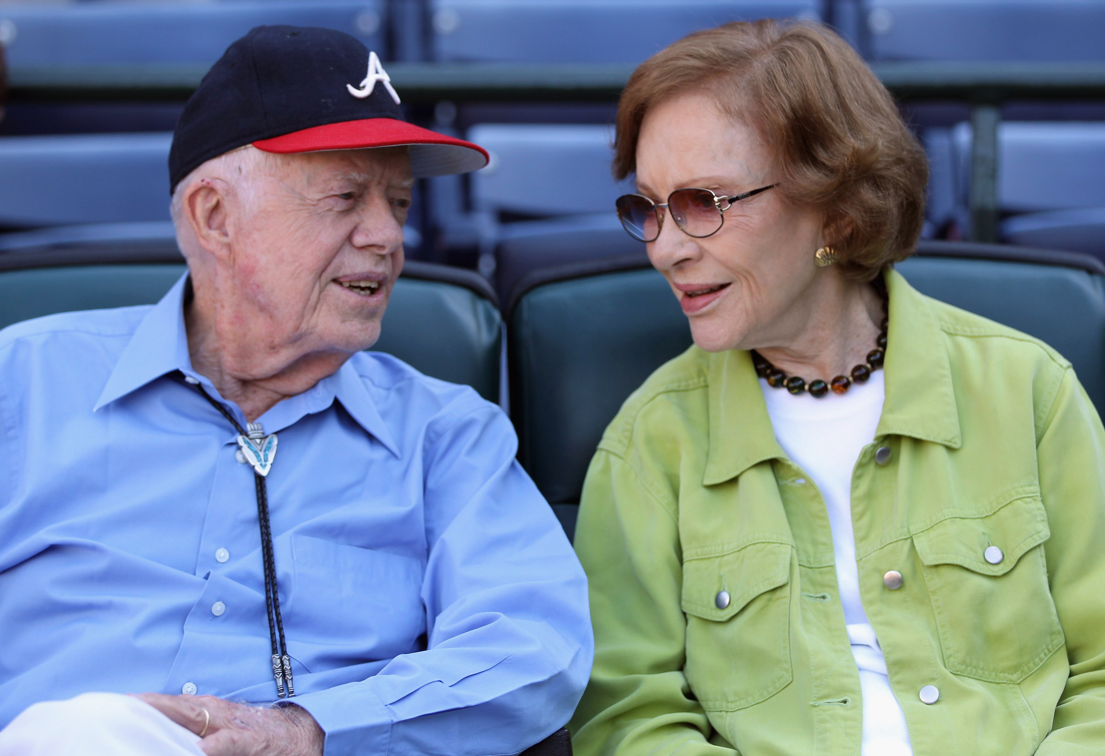 An elderly man in a blue shirt and cap chats with a woman in a green jacket and sunglasses. They sit closely, smiling at each other.