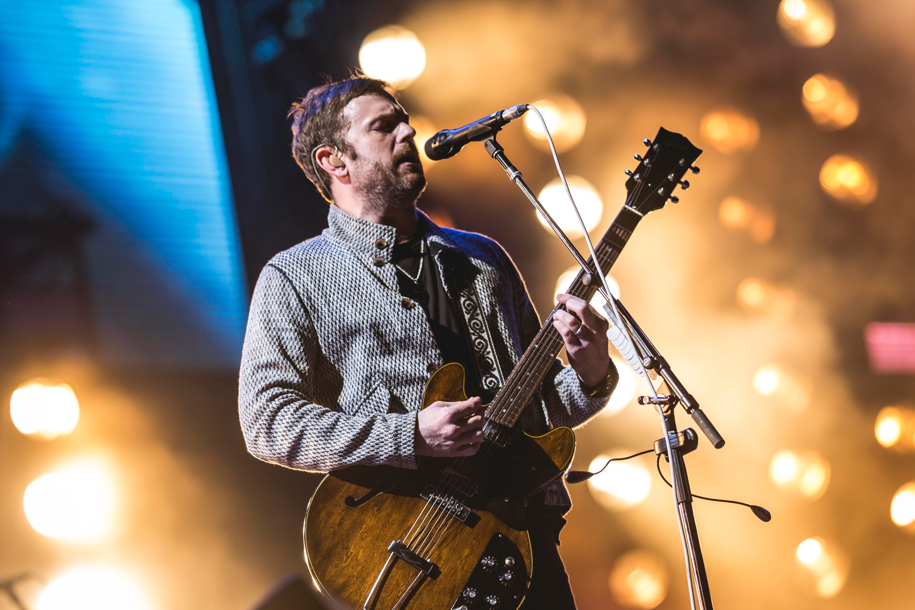 A man is playing an electric guitar and singing into a microphone on stage. He's wearing a patterned jacket, with bright stage lights glowing behind him.