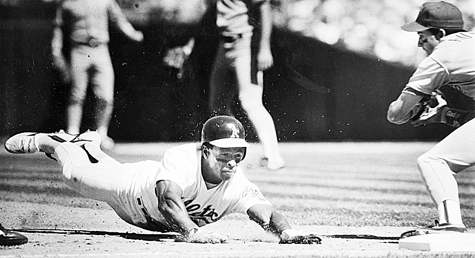 A baseball player wearing a helmet dives toward a base, stretching out his arms to reach it. Another player's legs are visible nearby, ready to tag him.