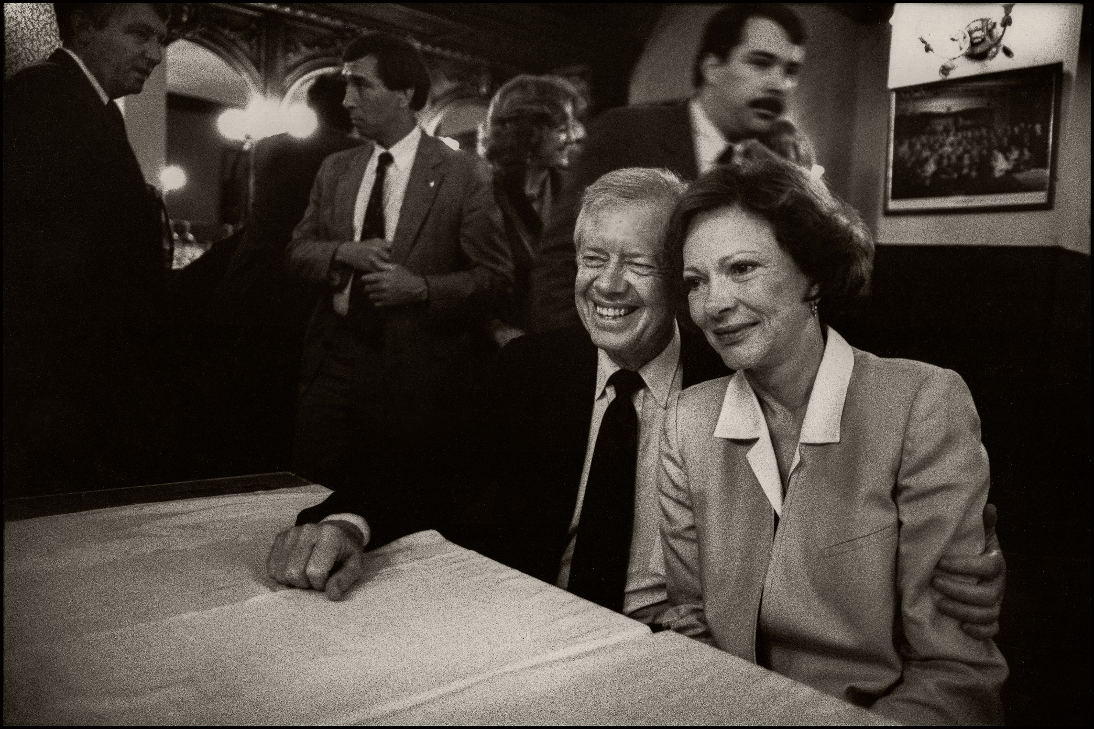 A smiling couple sits closely at a table, while several people stand in the background of a dimly lit room with ornate decorations.