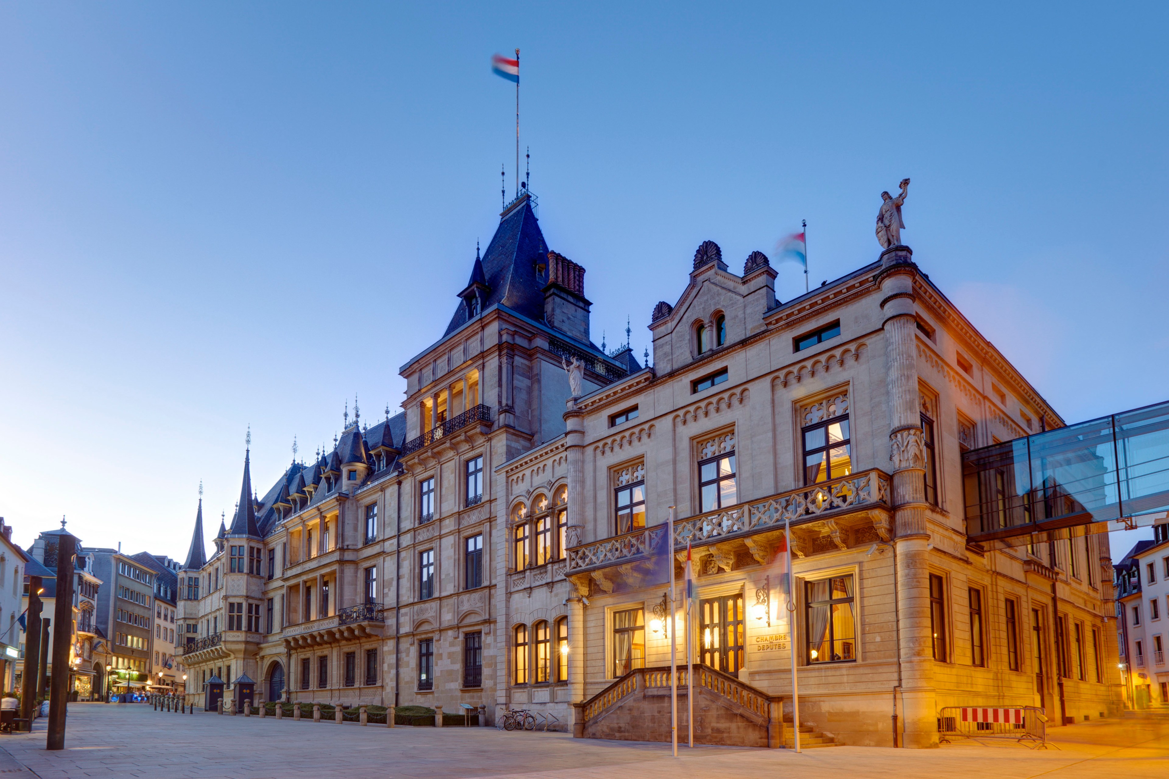 The image shows a grand historic building with ornate architectural details, peaked roofs, and flags. It's illuminated against a twilight sky, creating a majestic scene.