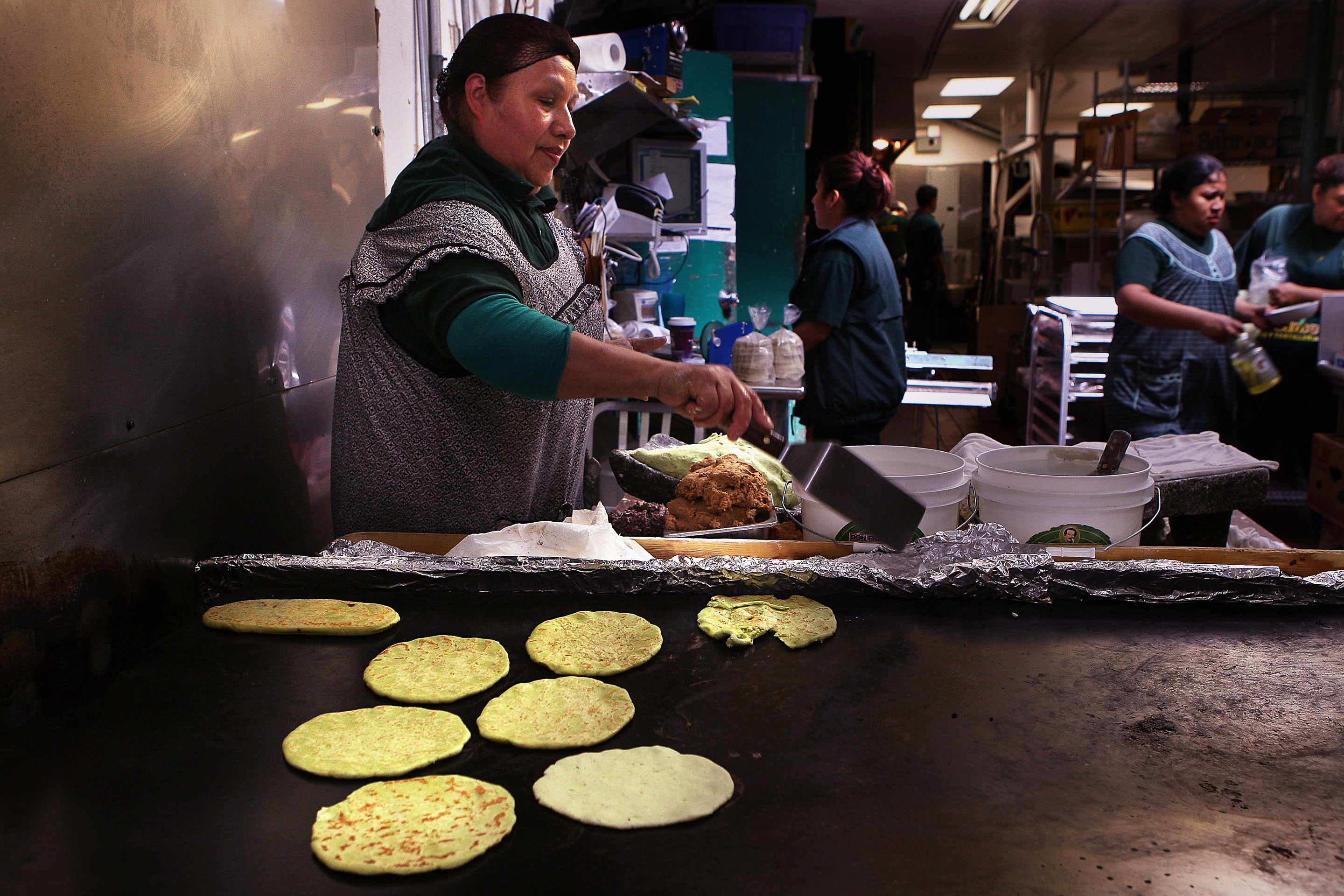 A woman in an apron cooks tortillas on a large griddle in a kitchen. Several people are in the background, and containers are nearby.