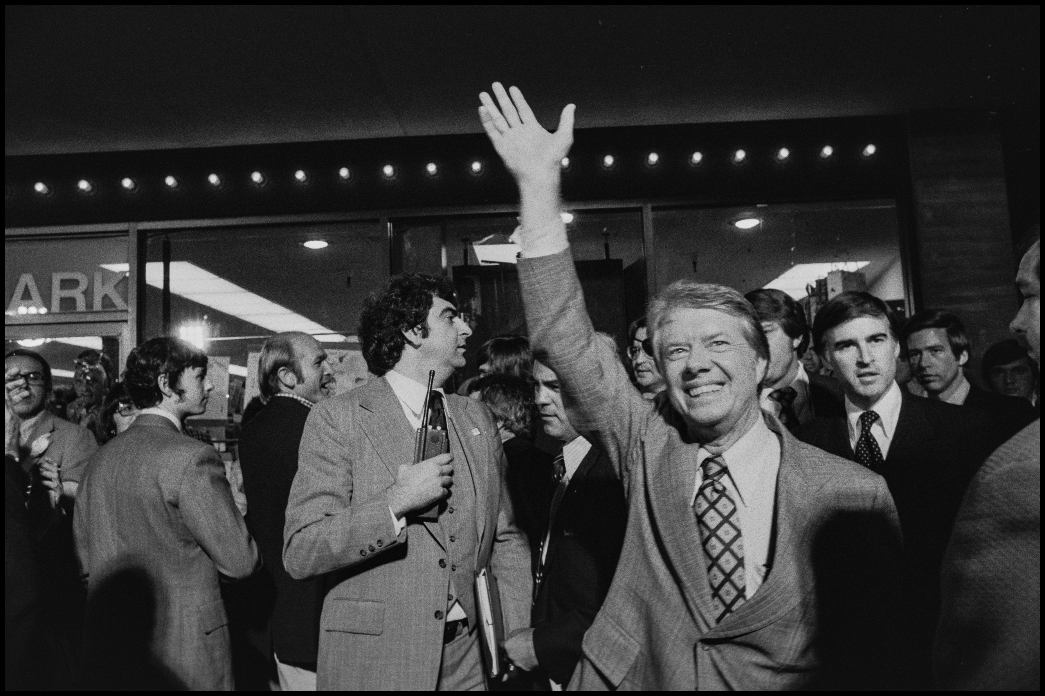 A smiling man in a suit waves to a crowd at an event, surrounded by other formally dressed individuals. The scene is lively with dim overhead lighting.