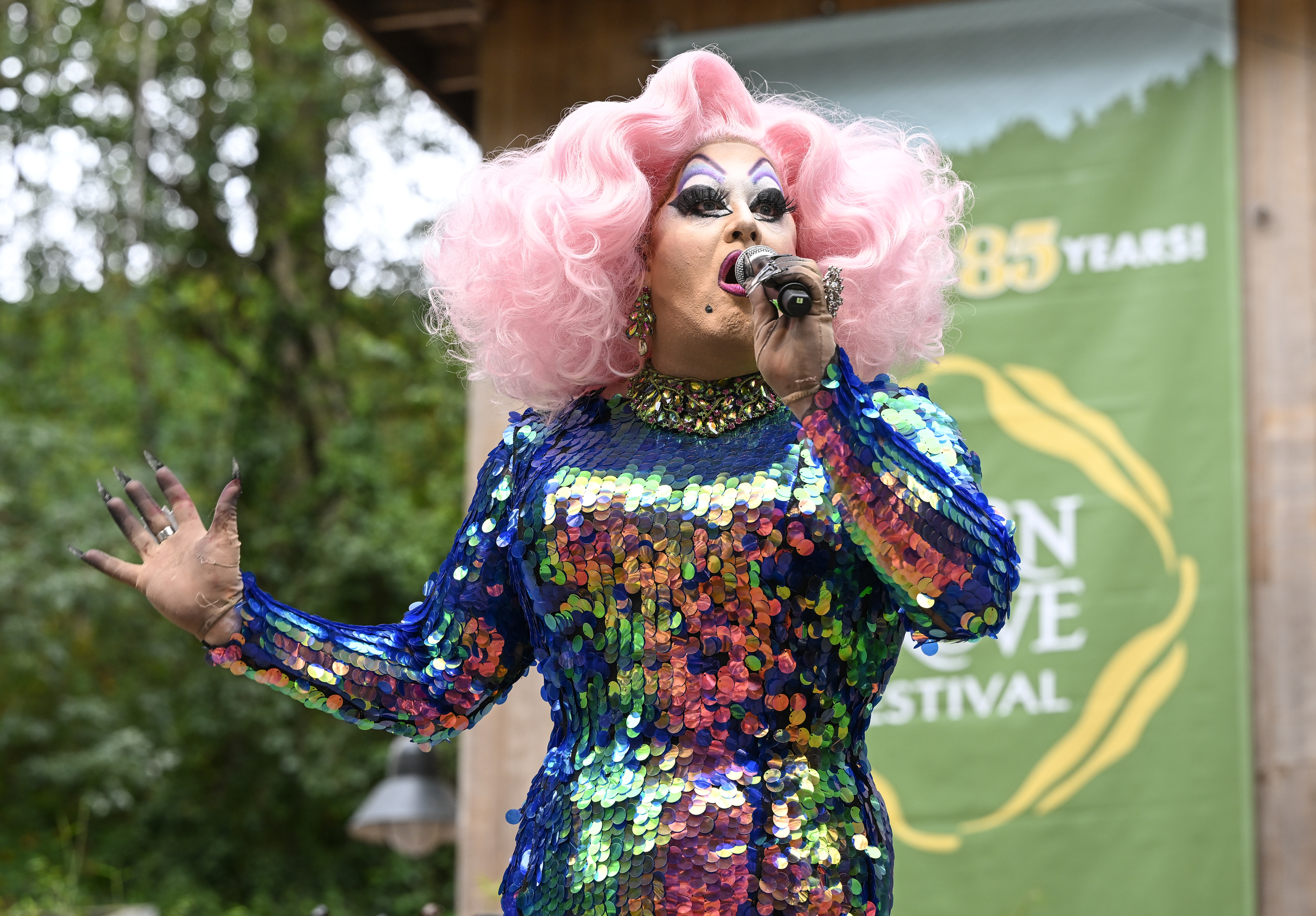 A performer in a sparkling, multicolored sequin dress sings into a microphone. They have dramatic makeup and voluminous pink hair, set against an outdoor festival backdrop.