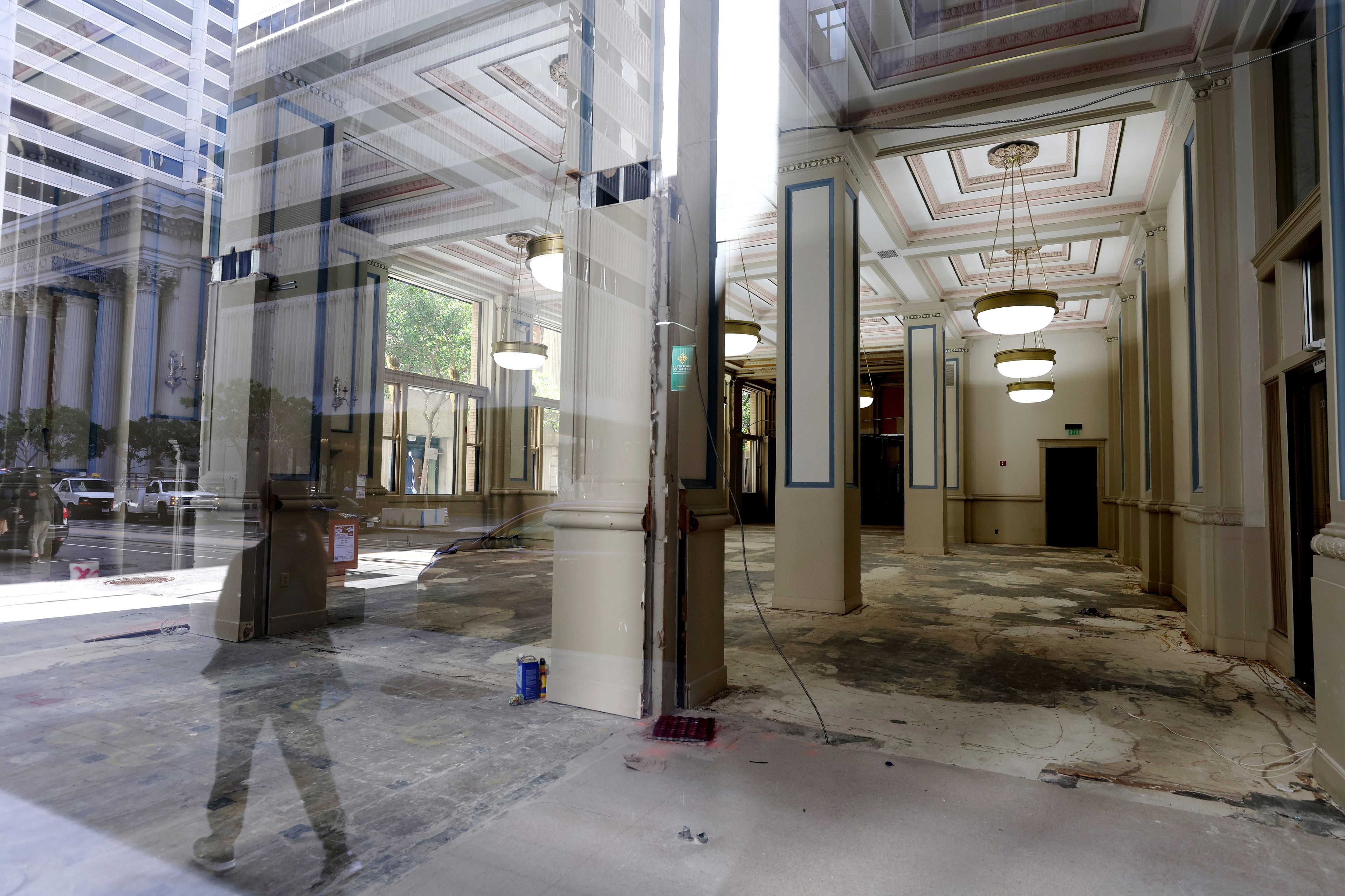 The image shows a large, empty interior space with high ceilings, ornate light fixtures, and visible floor damage. A reflection reveals a street scene outside.