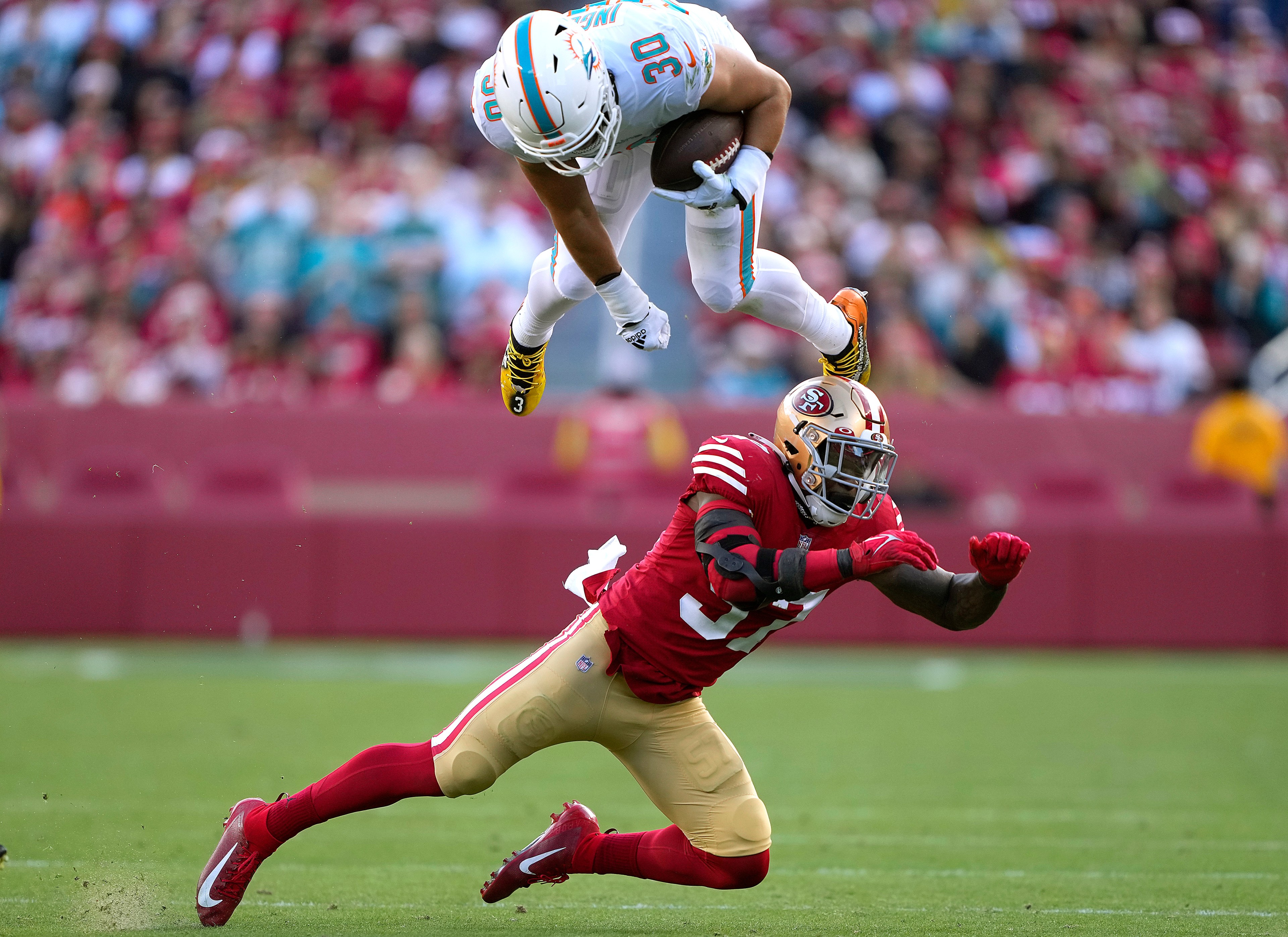 A football player in a white and teal uniform leaps over an opponent in a red and gold uniform during a game, with a blurred crowd in the background.