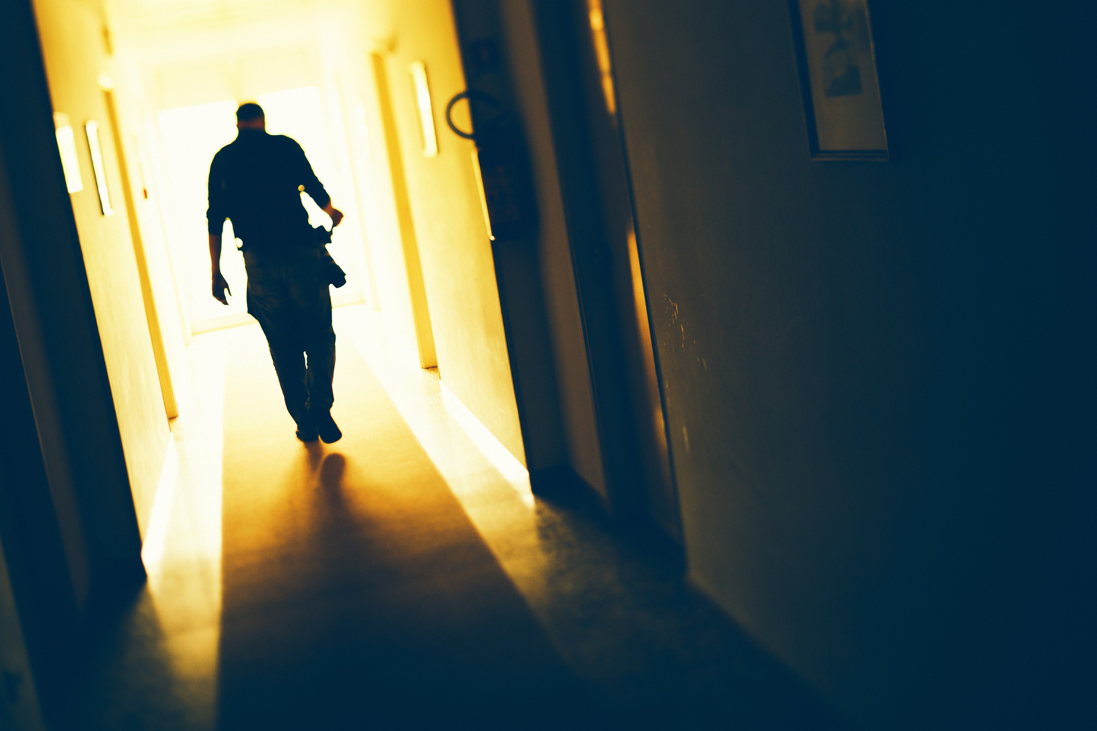 A silhouette of a person walks down a dimly lit hallway toward a bright doorway, casting a shadow on the floor. The scene is warm with golden hues.