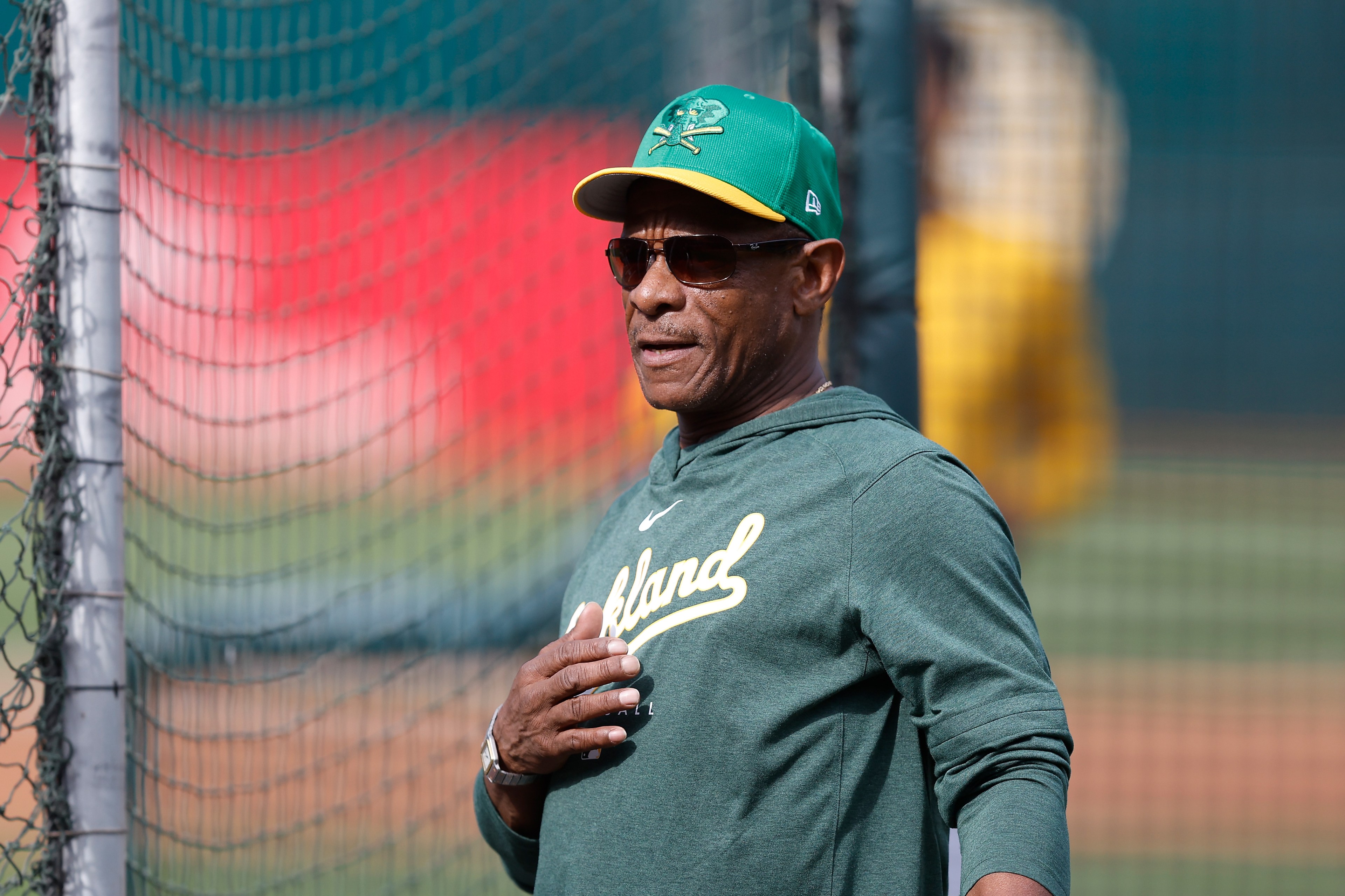 A person in an "Oakland" sweatshirt and green cap stands near a baseball net, wearing sunglasses and appearing relaxed, with one hand on their chest.