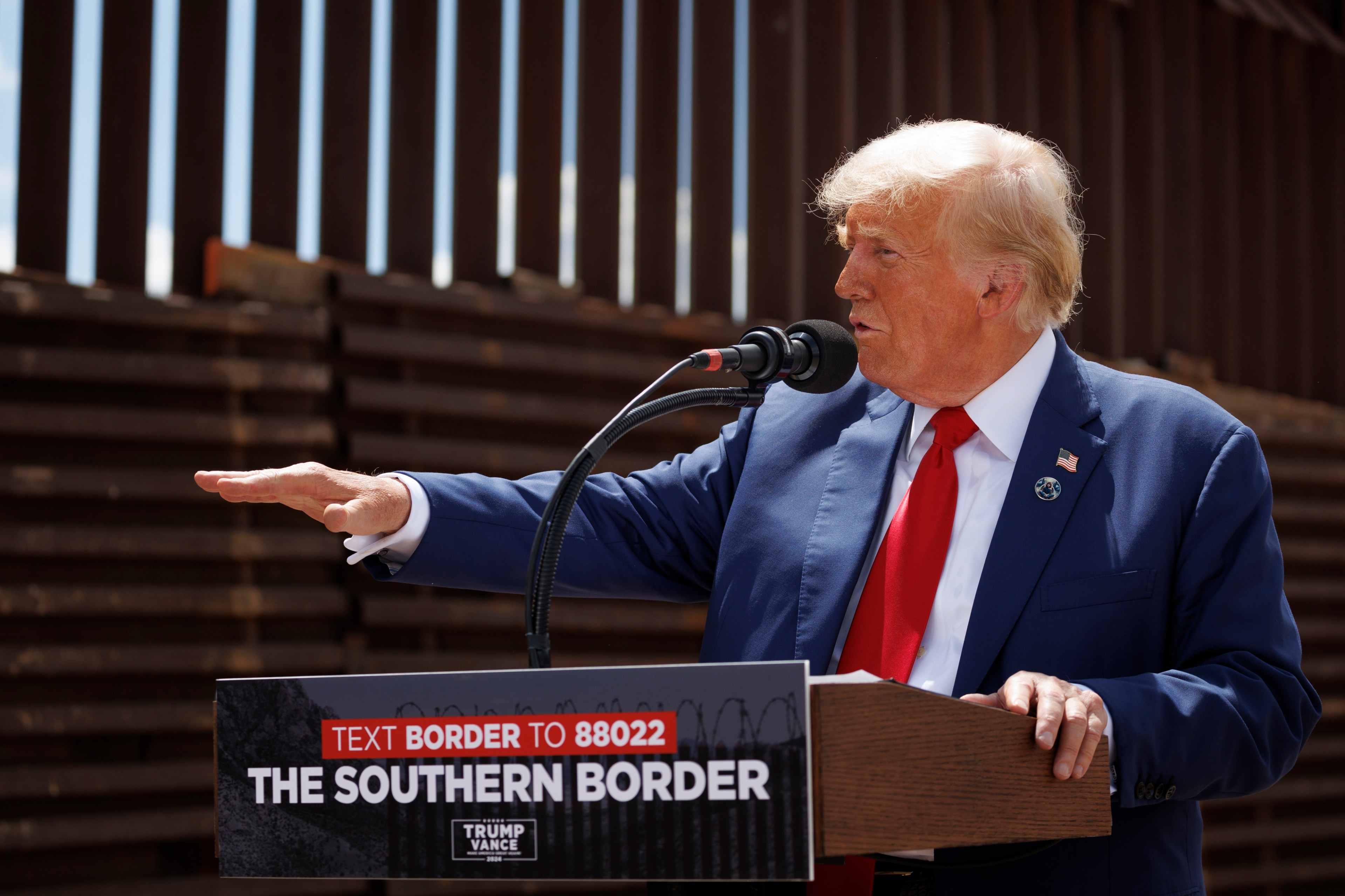 A man in a blue suit and red tie speaks at a podium with &quot;The Southern Border&quot; written on it, in front of a tall metal border fence.