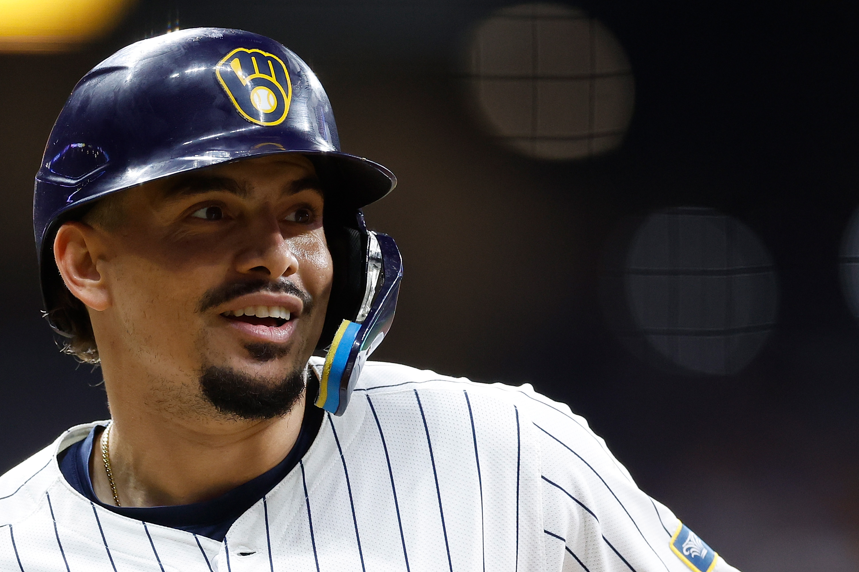 A baseball player smiles, wearing a dark helmet with a logo and a pinstriped jersey. The background is out of focus with circular lights.