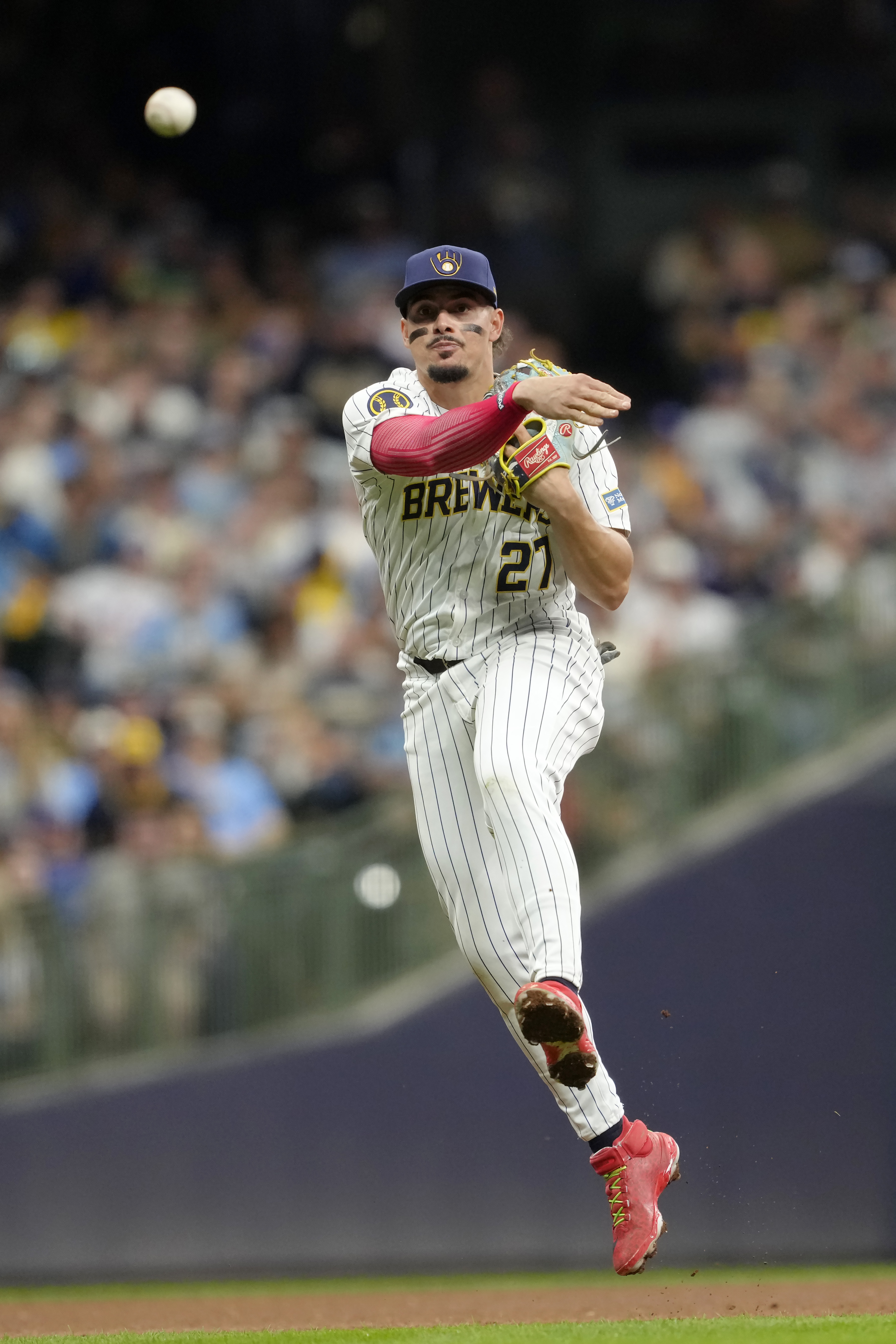 A baseball player in a Brewers uniform is in mid-action, throwing a ball. He wears a cap and red cleats, with a focused expression and a blurred crowd behind.