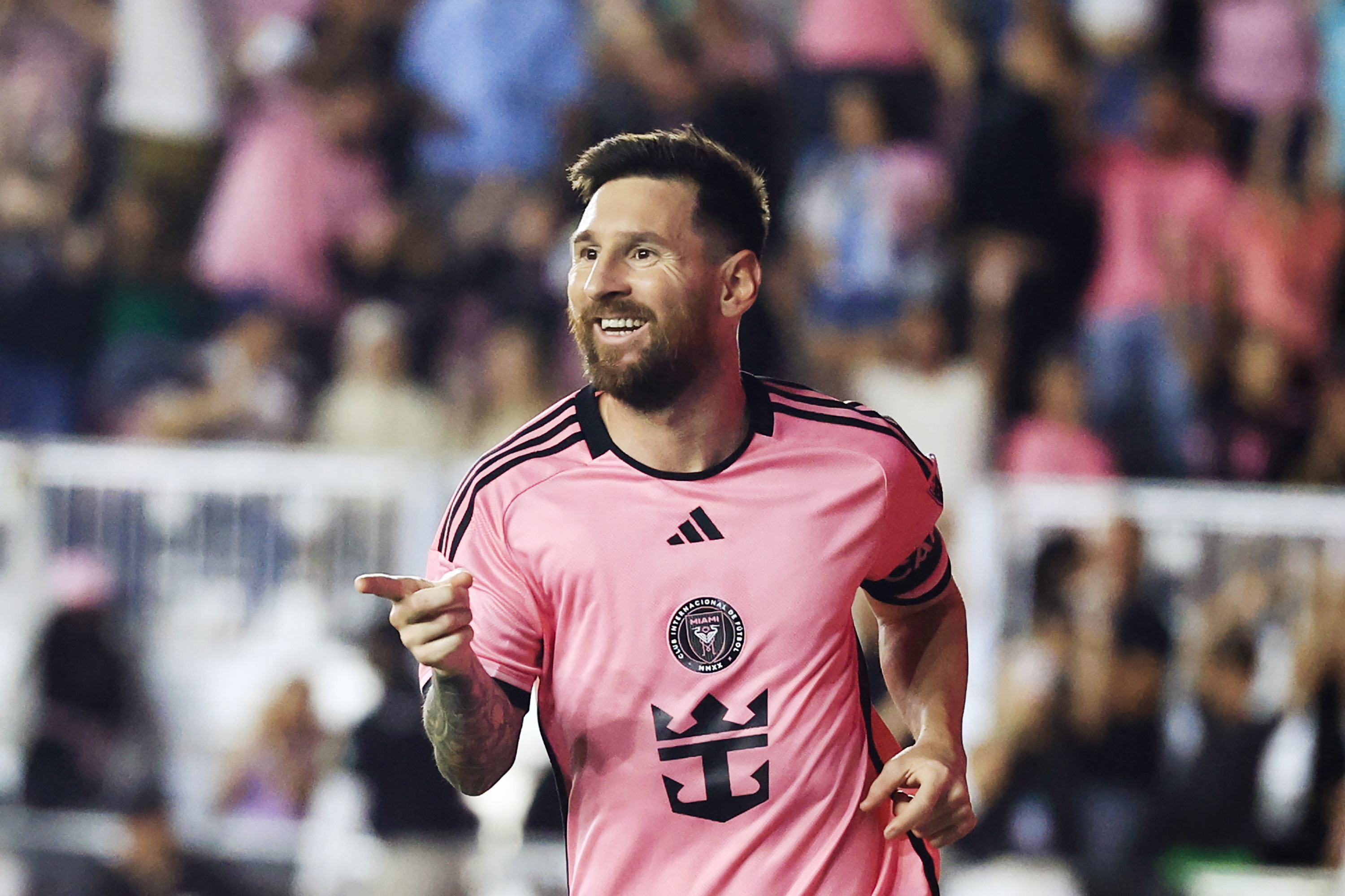 A soccer player in a pink jersey is smiling and pointing, with an enthusiastic crowd in the background.