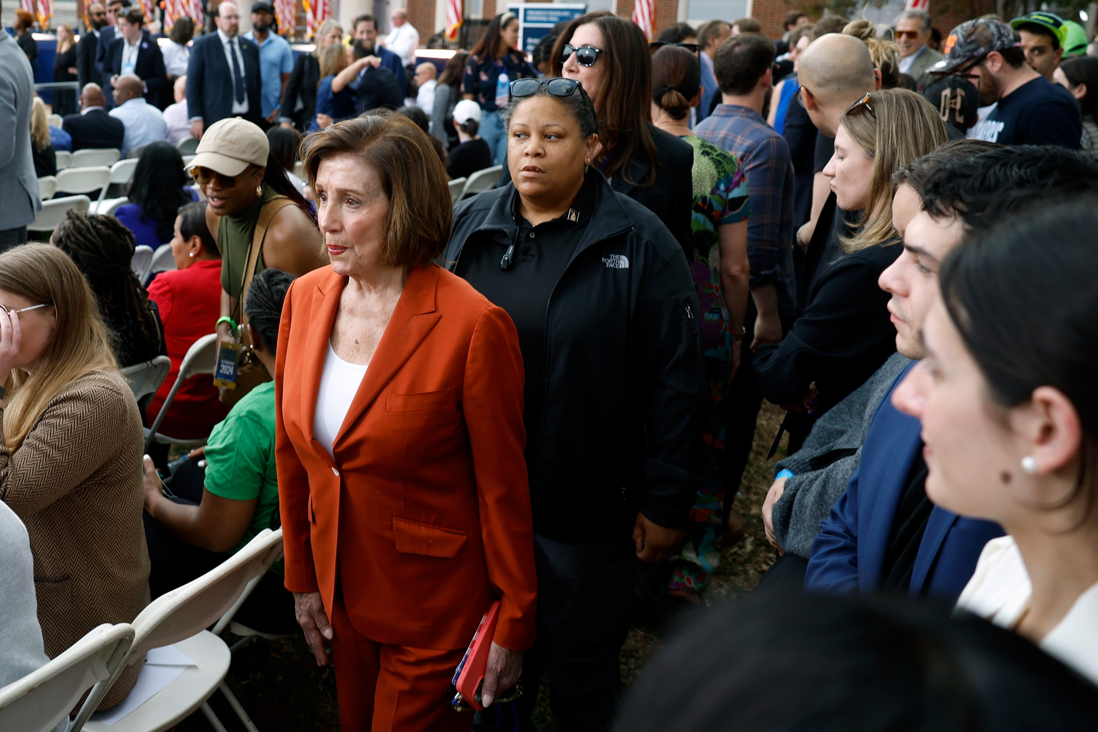 A woman in an orange suit stands amidst a crowd seated outdoors, with people talking and walking around. In the background, there are American flags.