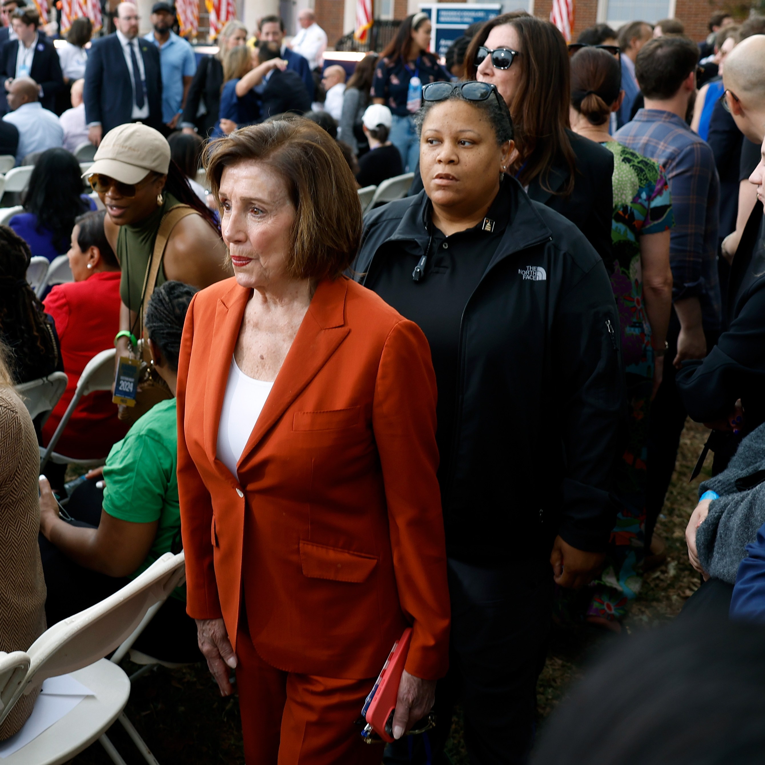 A woman in an orange suit stands amidst a crowd seated outdoors, with people talking and walking around. In the background, there are American flags.