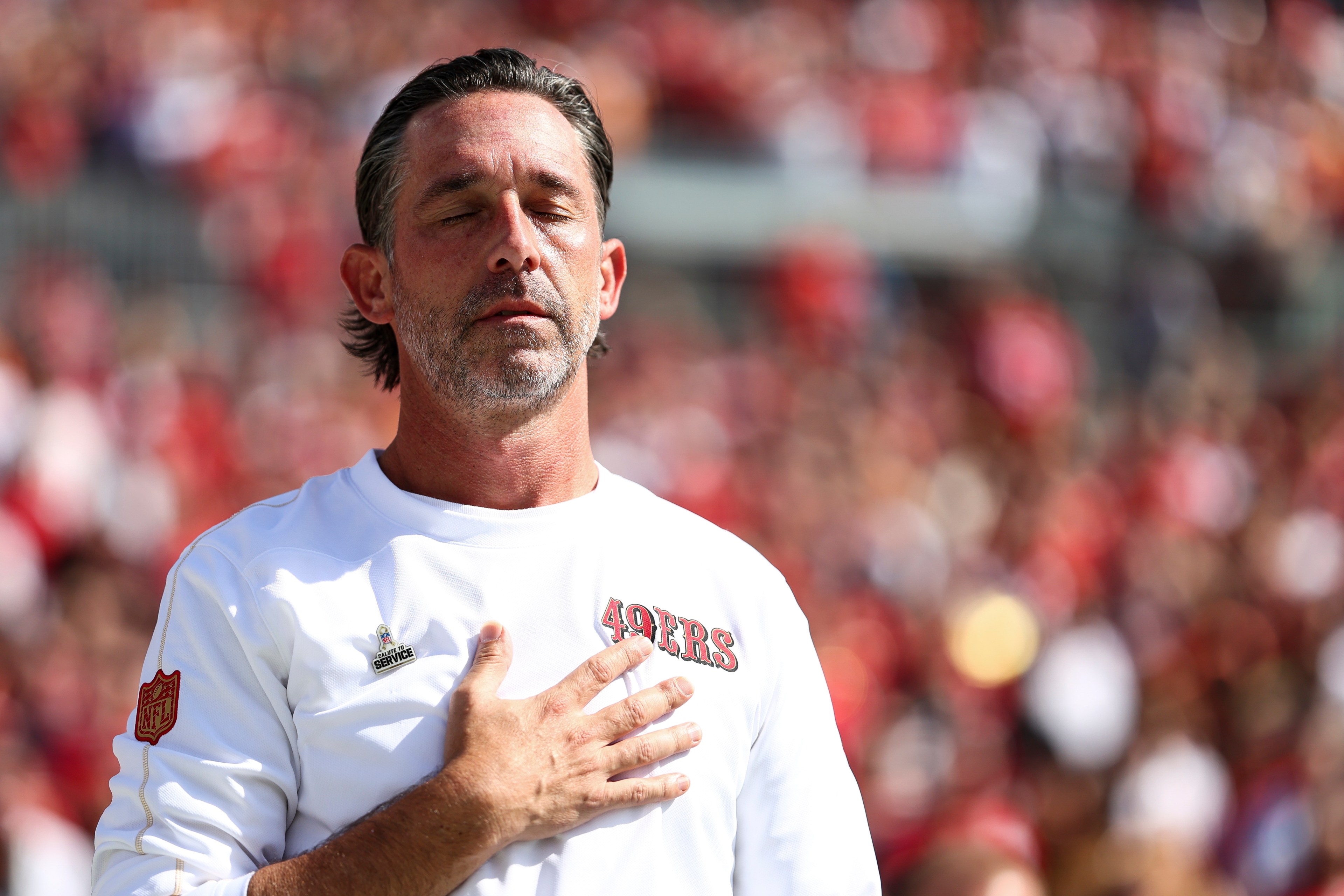 A man in a 49ers shirt stands with his eyes closed, hand on his chest, against a blurred crowd background.