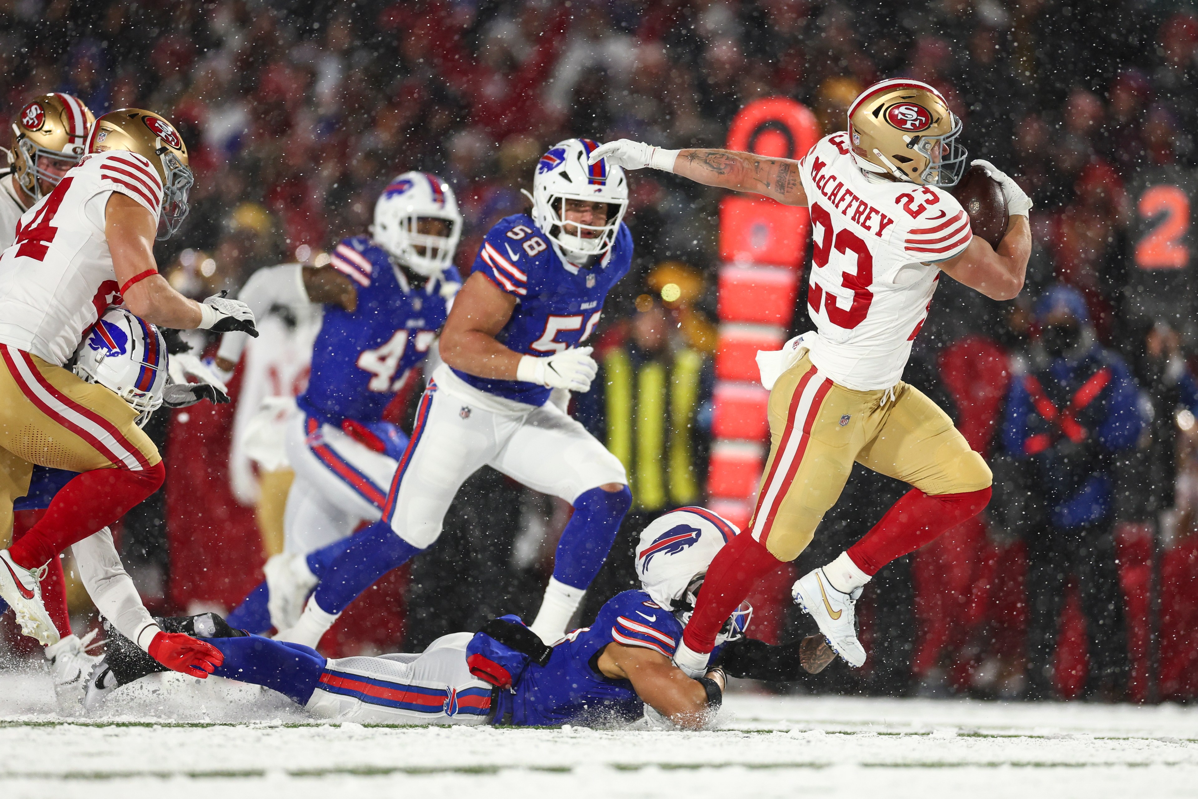 A football player in a red and gold uniform runs with the ball while avoiding a tackle on a snowy field. Other players in blue jerseys attempt to block him.