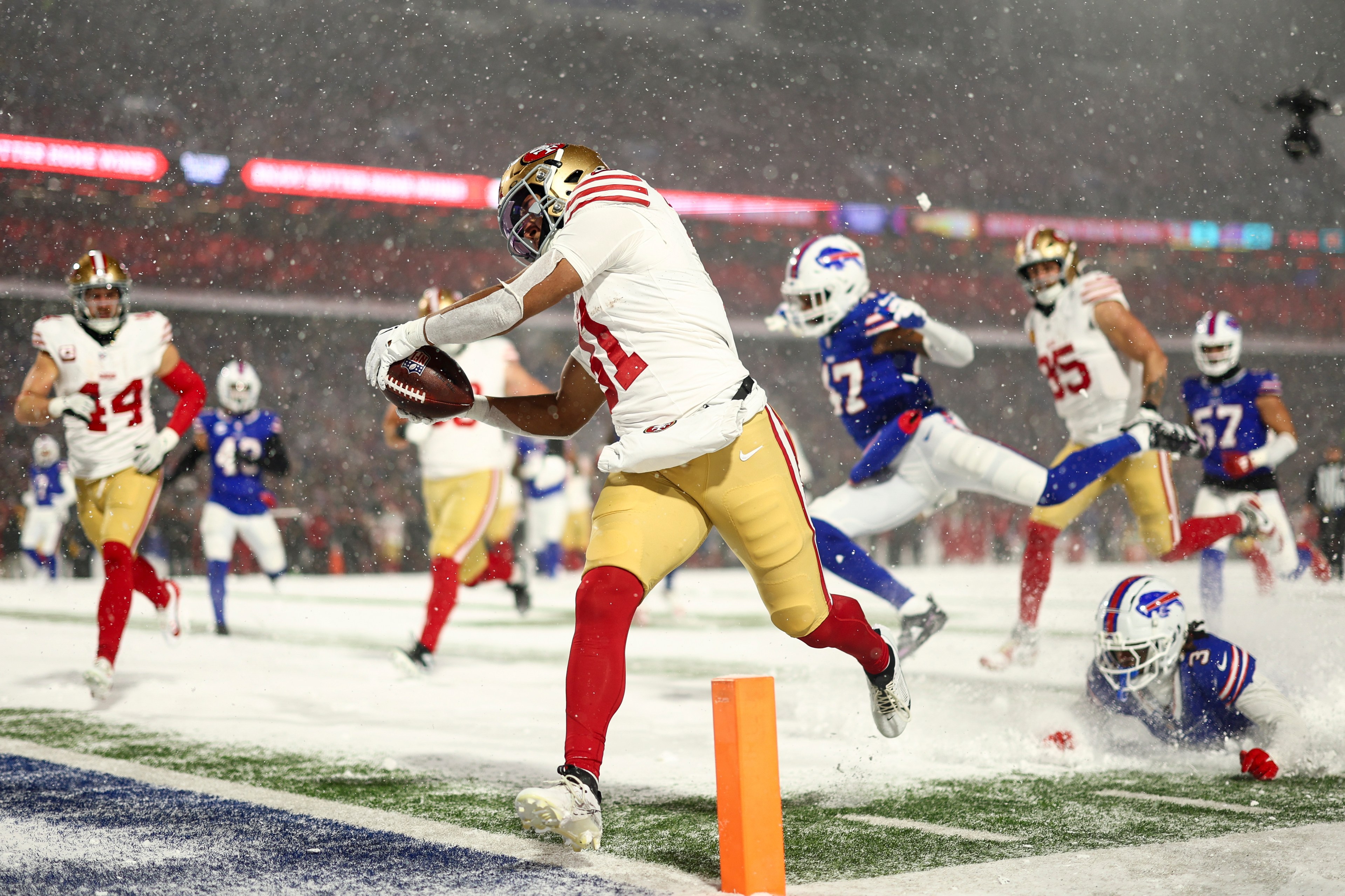 A football player in a white and red uniform runs into the end zone for a touchdown, with defenders in blue and white diving behind him on a snowy field.