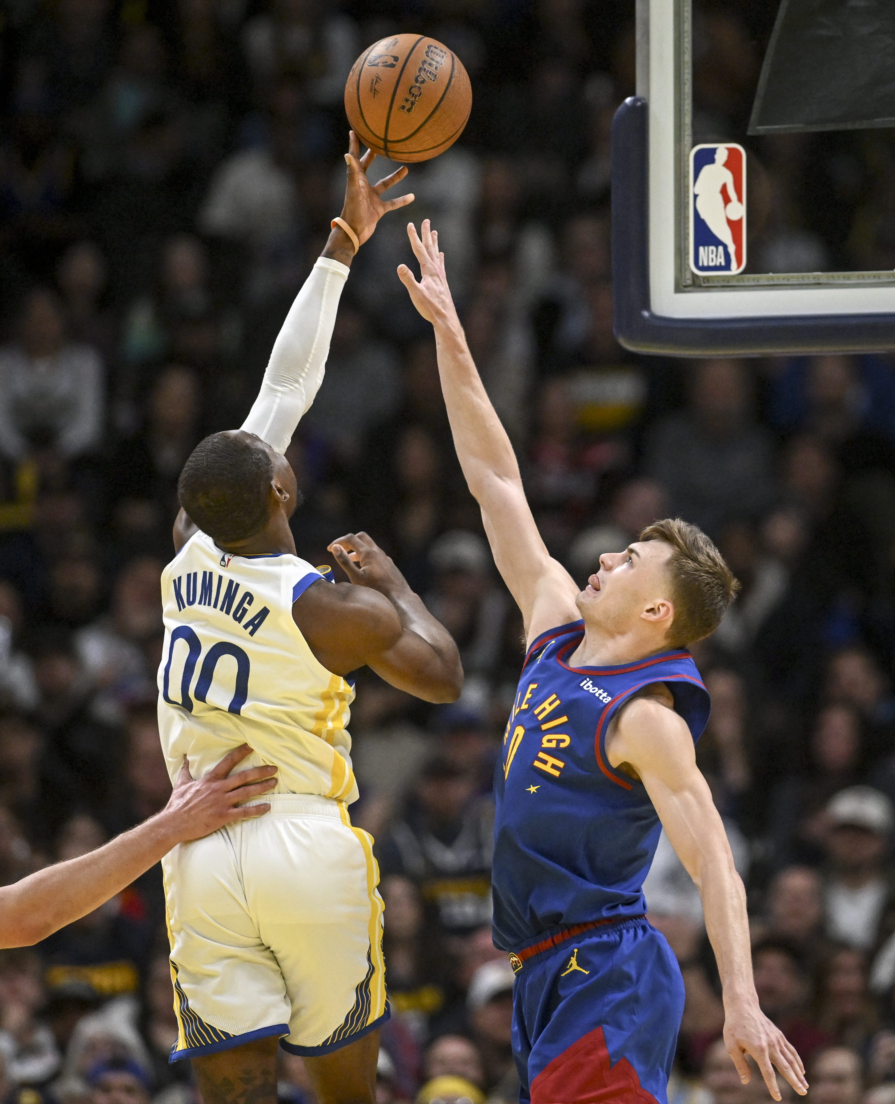 Two basketball players jump to reach the ball. One in a white jersey, named Kuminga, extends his arm while a player in a blue jersey tries to block him.