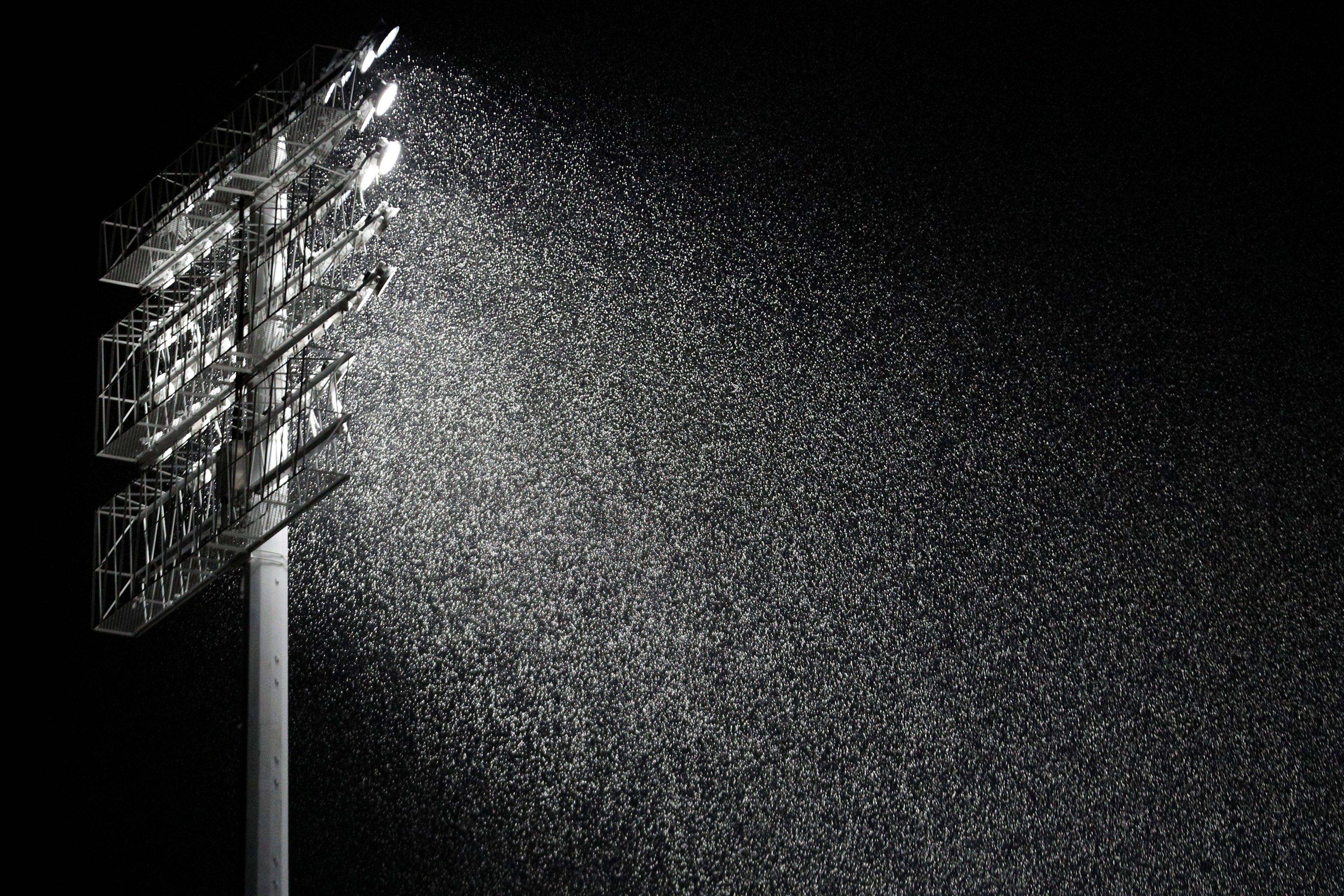 A tall stadium light beam shines through heavy rain in the dark, creating a bright, shimmering effect as raindrops are illuminated against the black background.