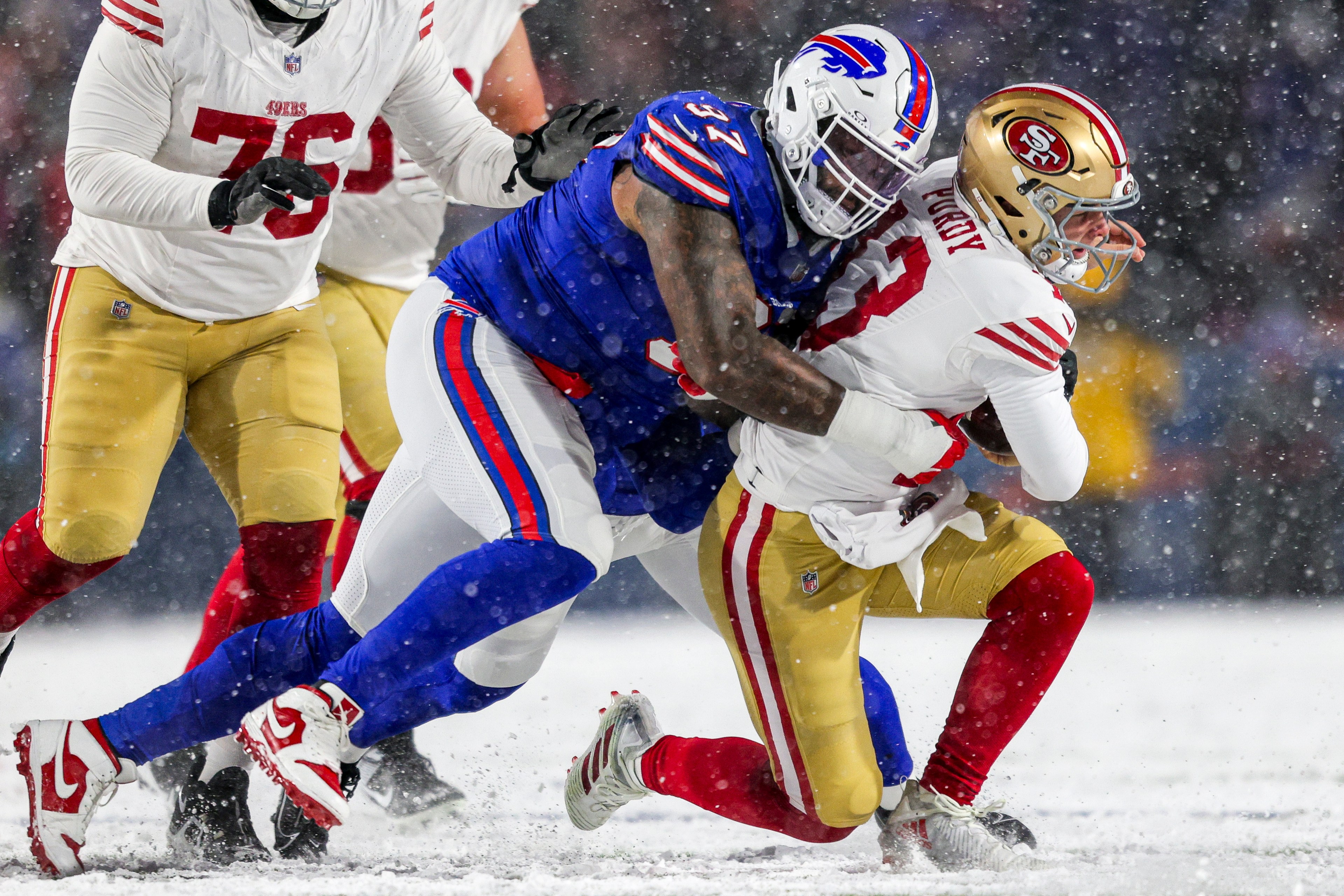 A football player in a blue and white uniform tackles another player in a red and gold uniform on a snow-covered field during a game.