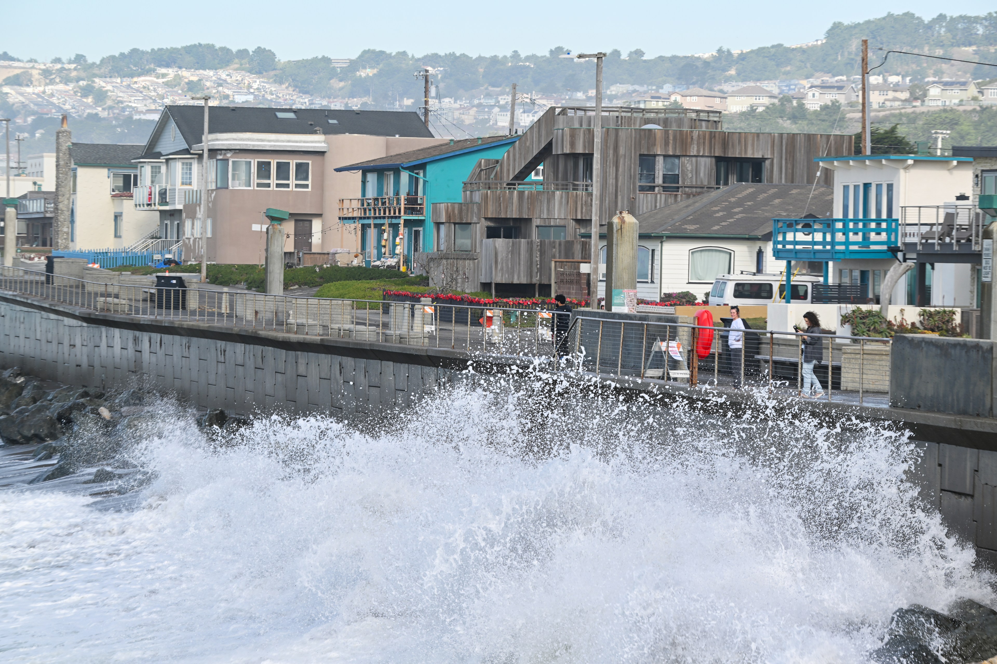 Waves crash against a seawall in front of colorful houses, while people walk along a pathway beside the sea. Hills and more buildings are visible in the background.