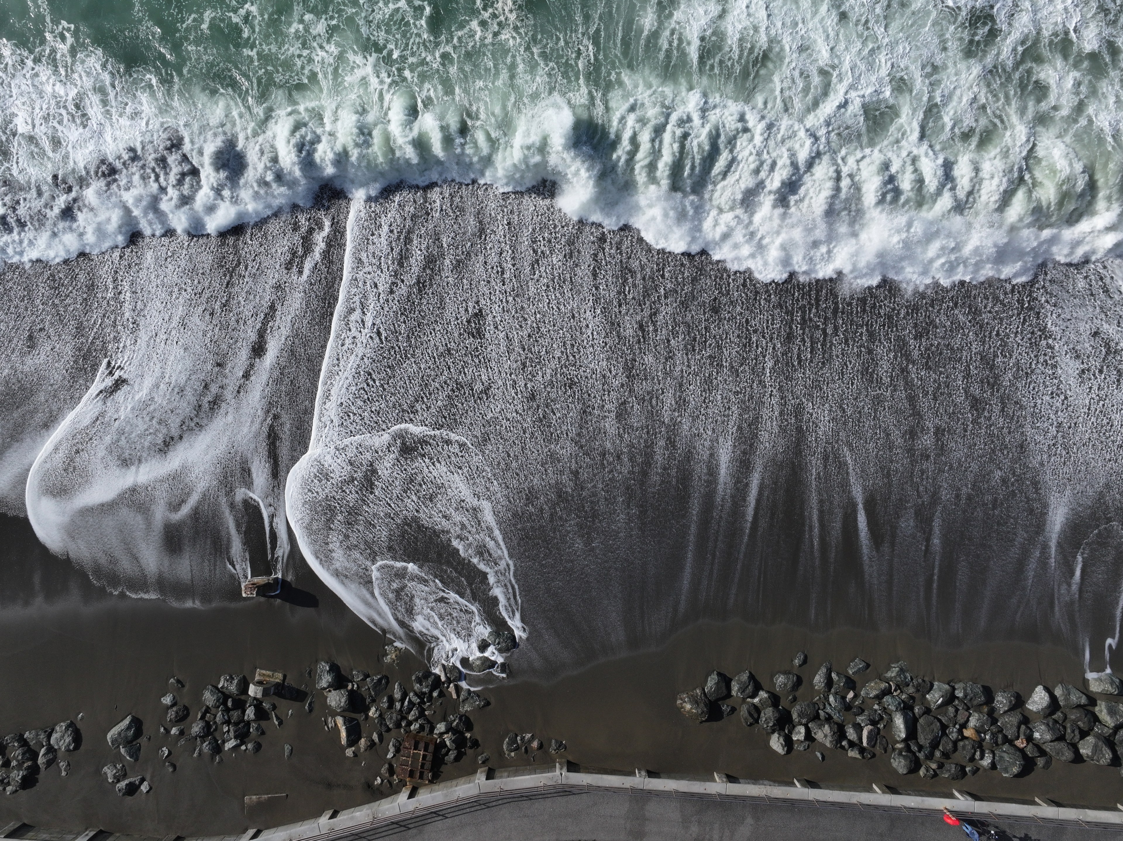 Waves crash onto a dark, rocky beach, spreading white foam across the sand. A row of large stones lines the shoreline, and a road runs along the bottom edge.