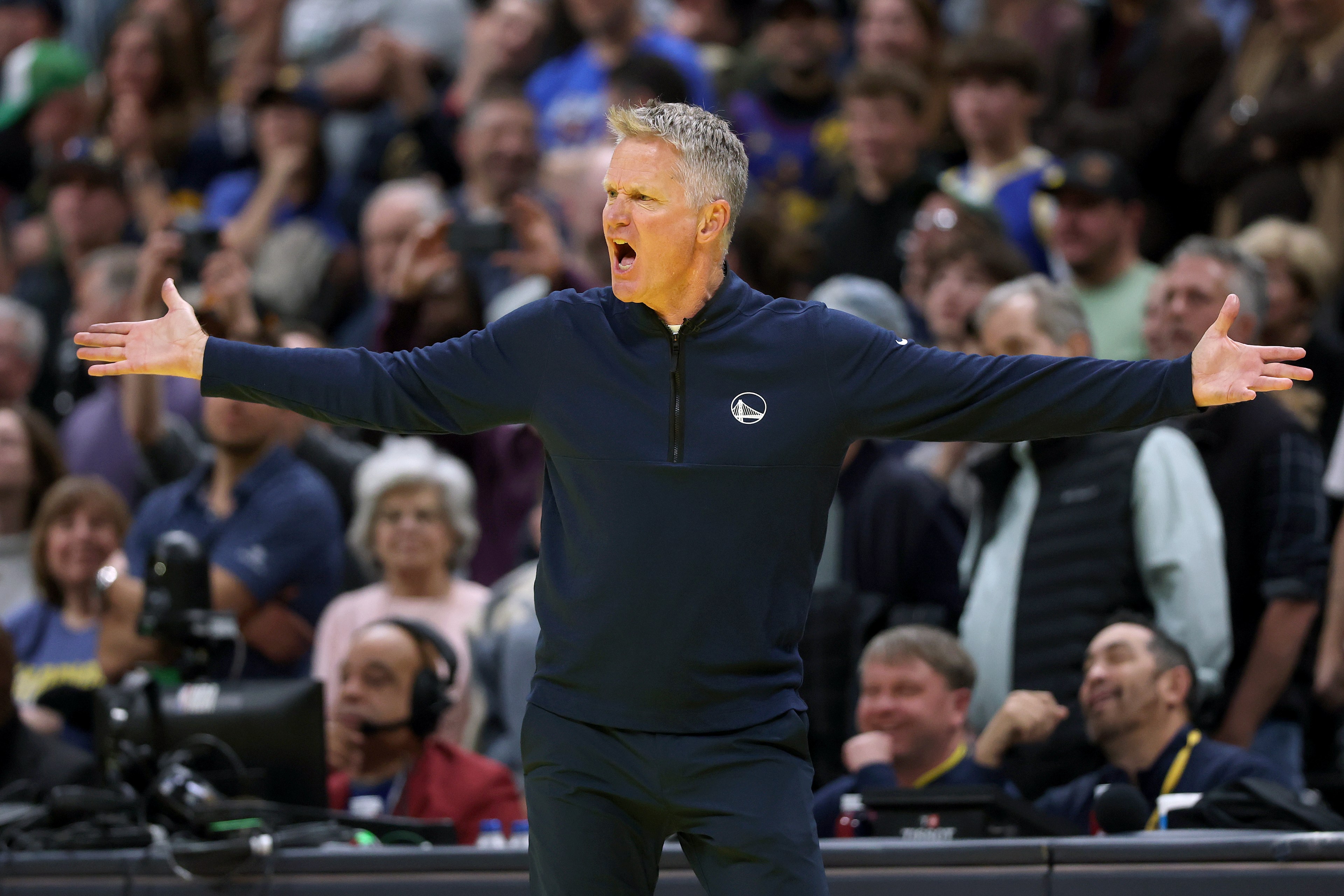 A basketball coach wearing a dark blue jacket with a logo is on the court, arms outstretched, appearing animated as he communicates during a game.