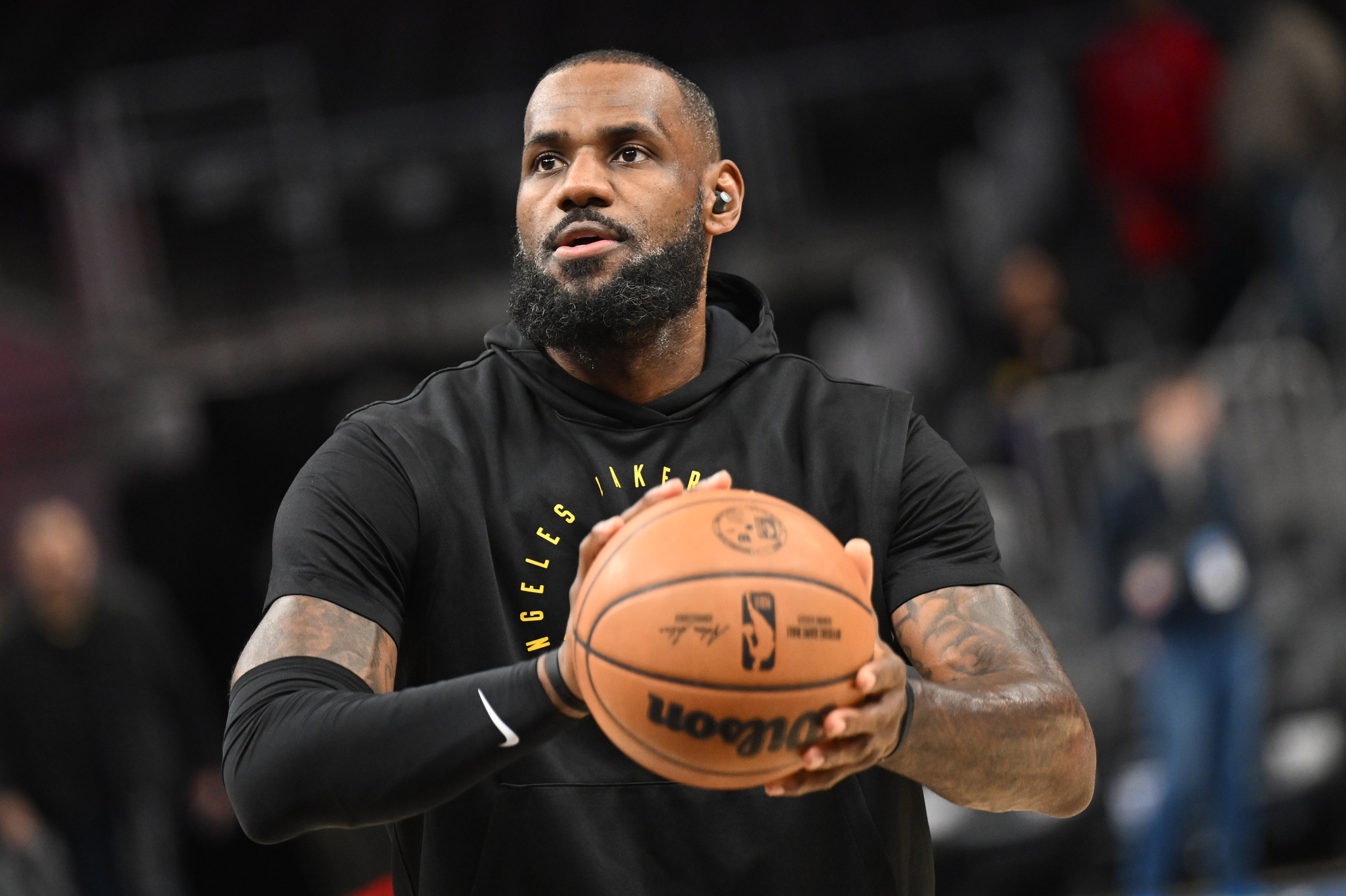 A man with a beard, wearing a black sports hoodie, is focused while holding a basketball. The background is blurred, suggesting an indoor sports venue.