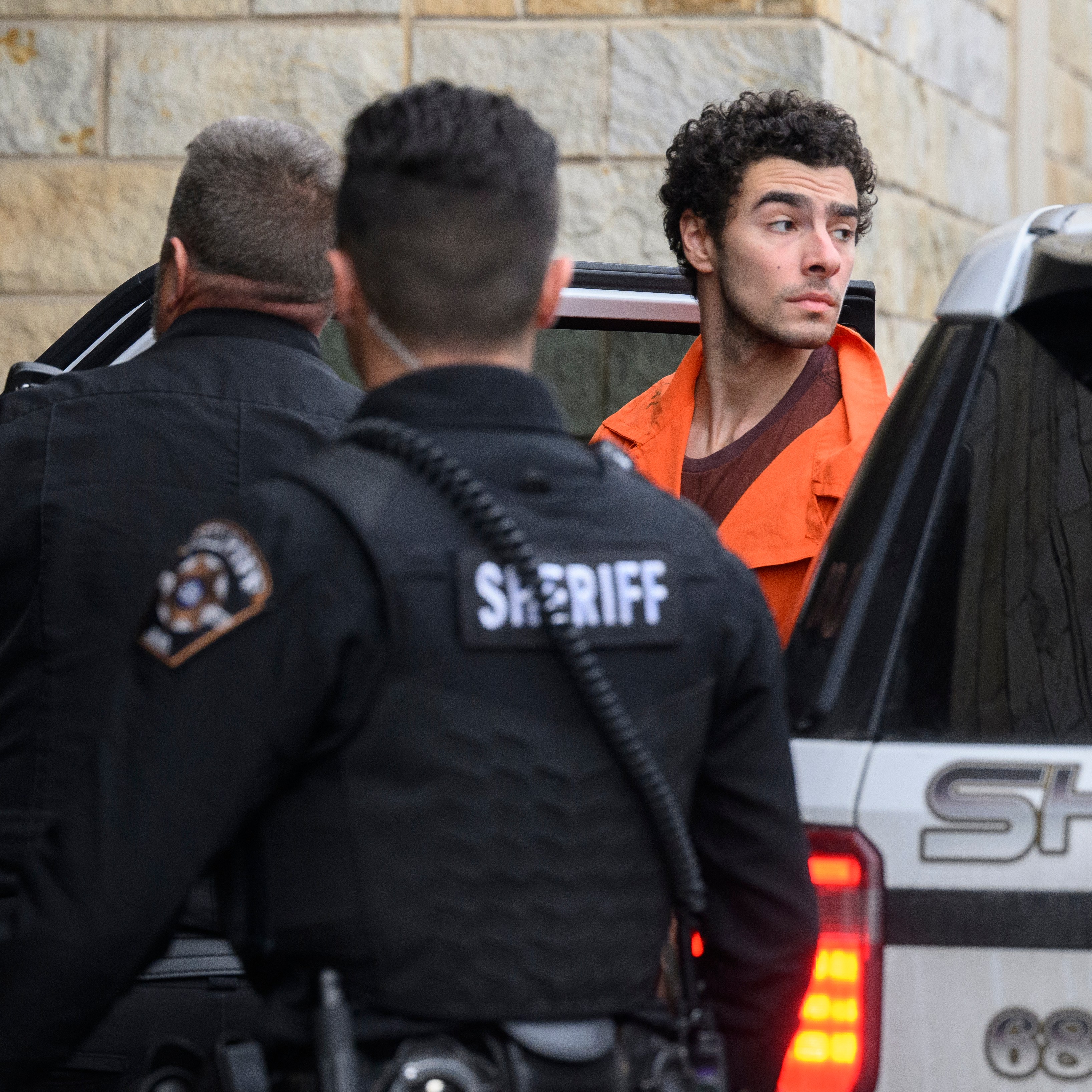 A man in an orange jumpsuit exits a sheriff's vehicle, accompanied by two officers in black uniforms. They are outside a stone building.