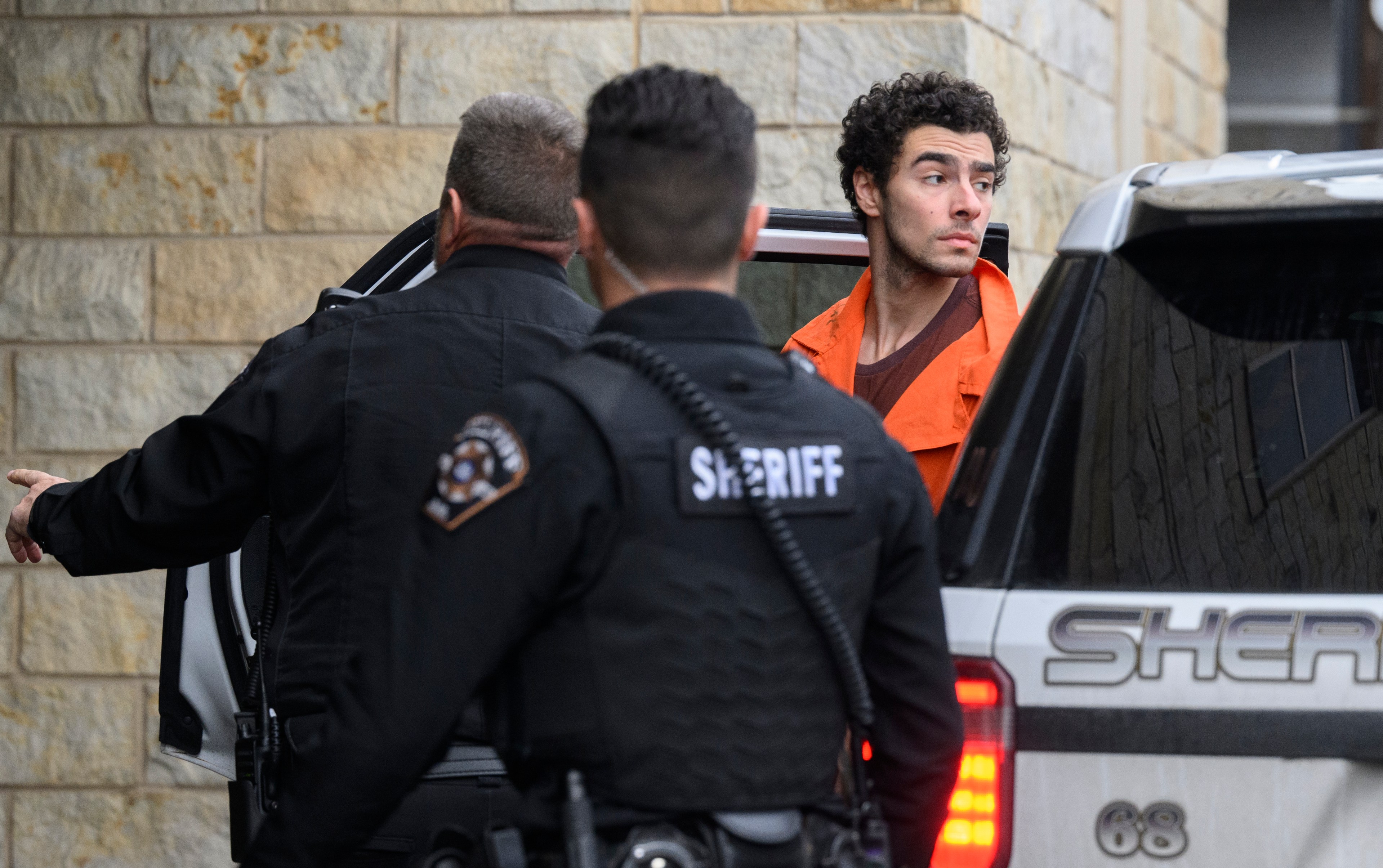 A man in an orange jumpsuit exits a sheriff's vehicle, accompanied by two officers in black uniforms. They are outside a stone building.