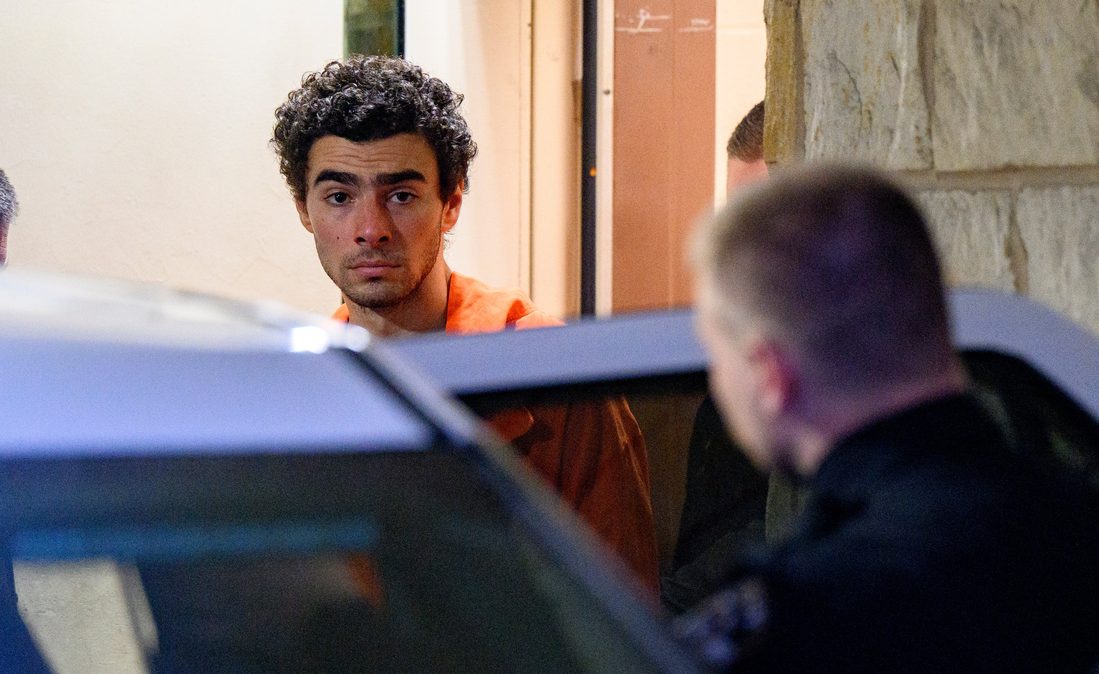 A man in an orange jumpsuit stares intently beside a vehicle, with an officer nearby, in a dimly lit setting.