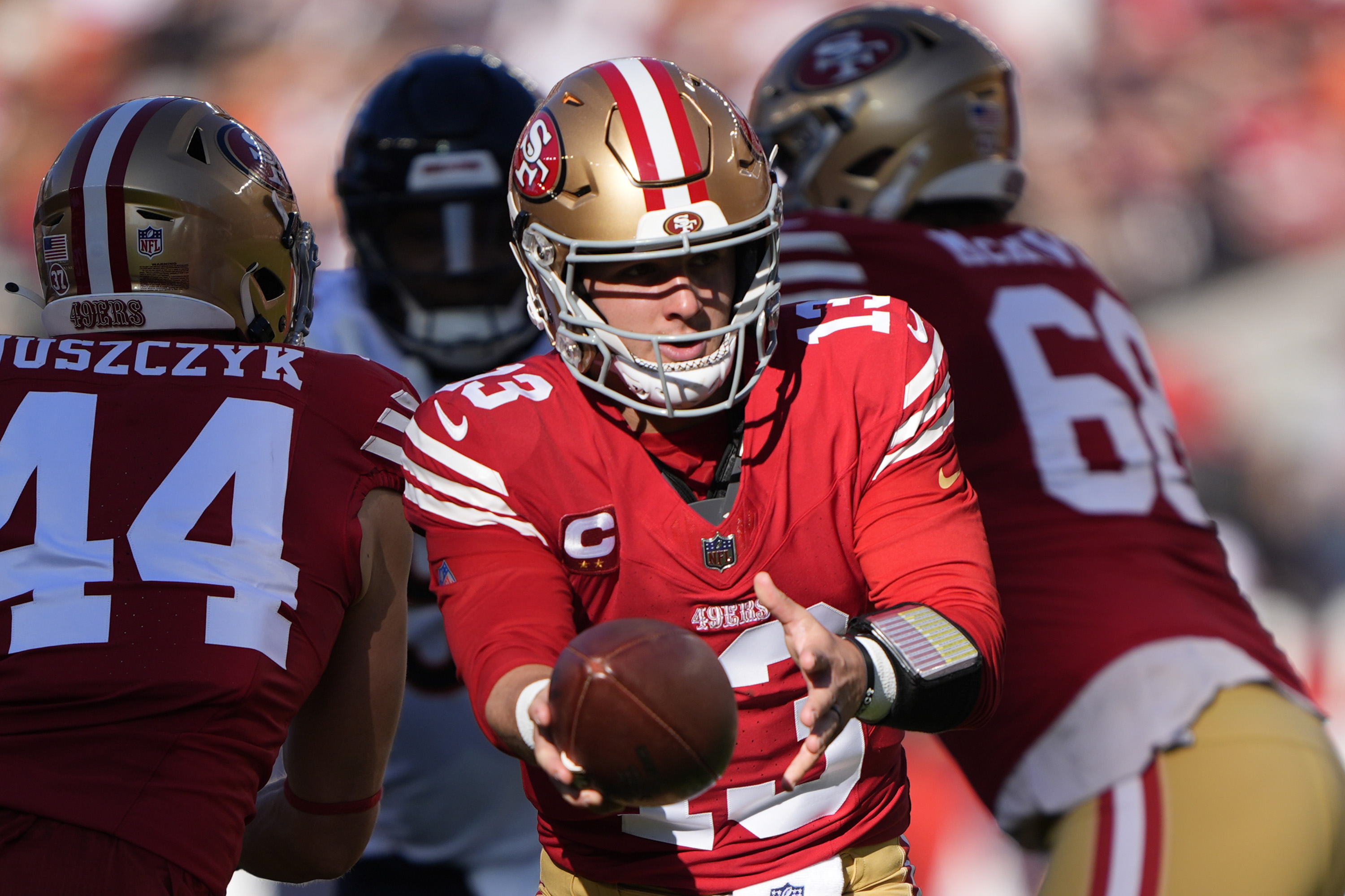 A football player in a red and gold uniform, wearing a helmet, is handing off the ball to a teammate. Another player is visible in the background.