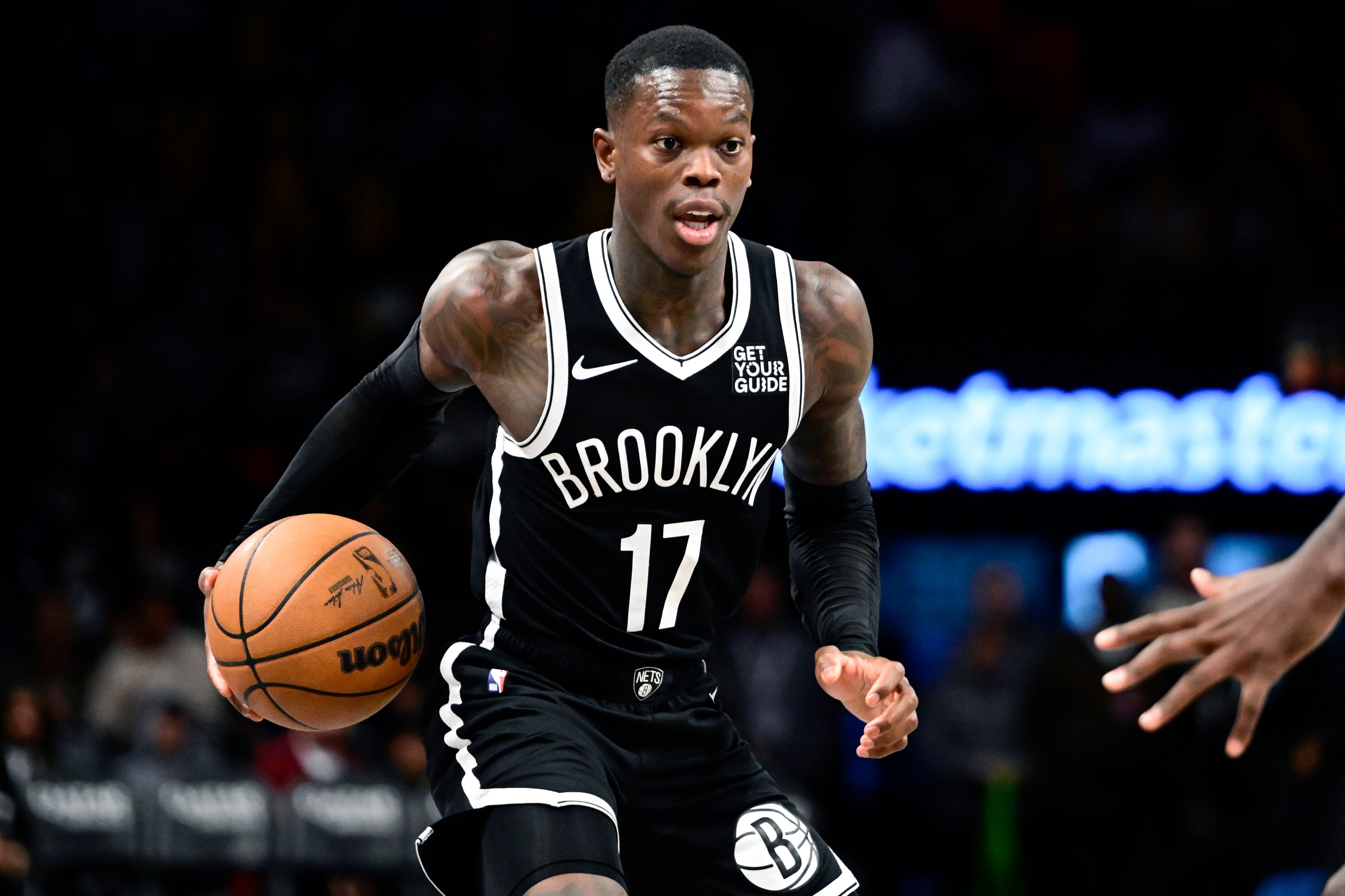 A basketball player in a black Brooklyn jersey, number 17, dribbles the ball on a court. He's focused and in action, with a blurred audience in the background.