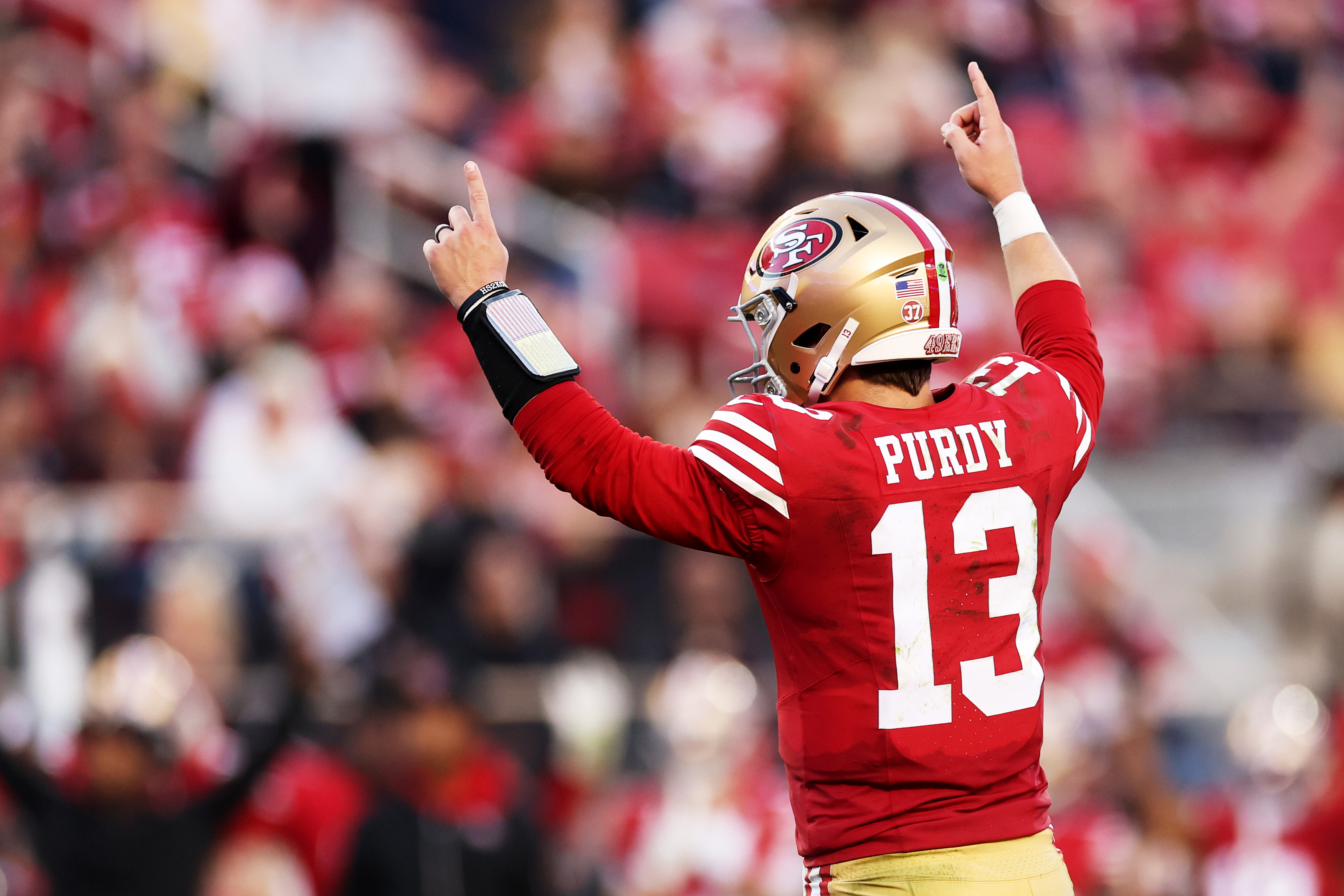 A football player in a red jersey with "Purdy" and number 13 raises his arms with index fingers pointed up, celebrating on a crowded field.