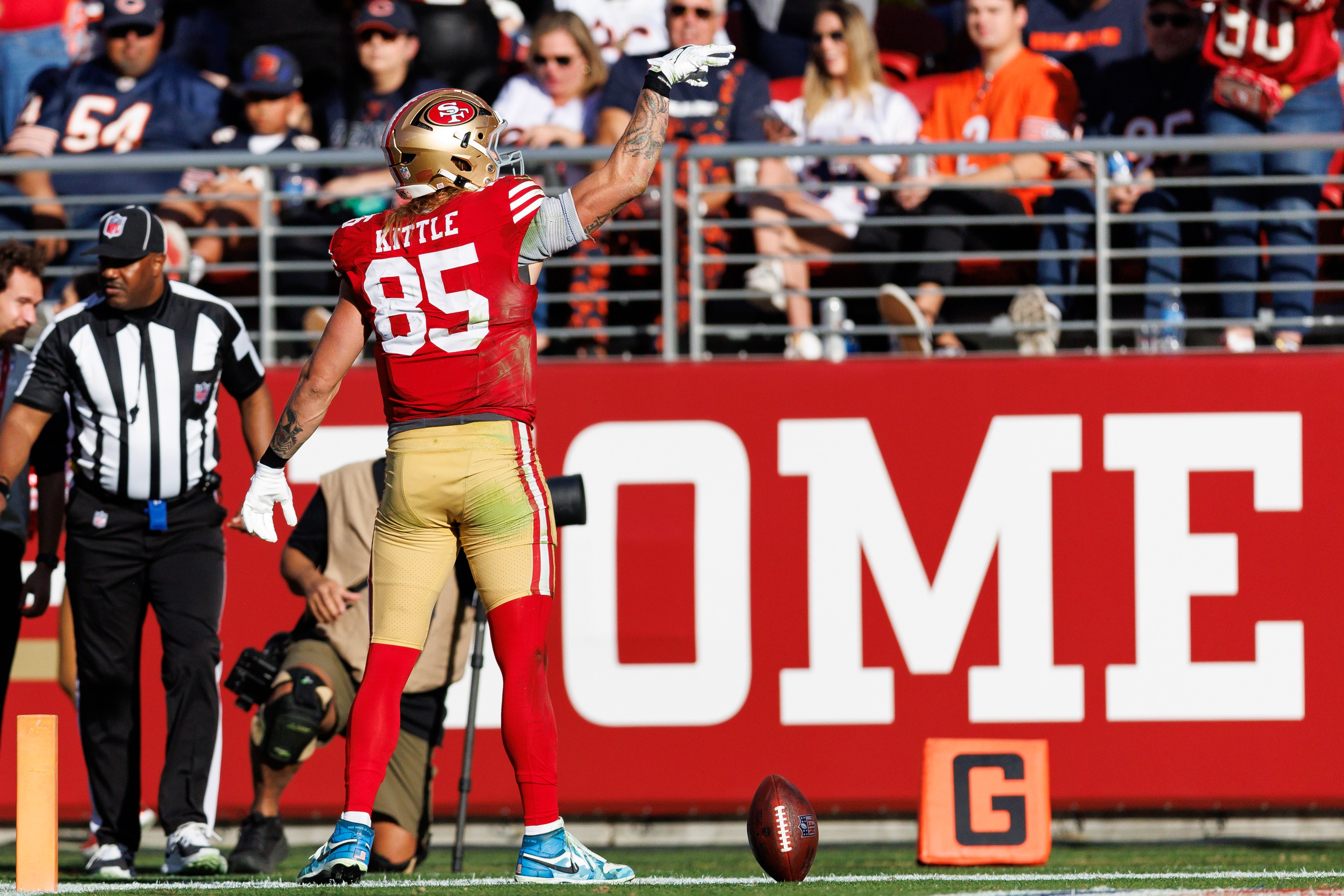 A football player in a red and gold uniform stands on the field gesturing. The number 85 is visible on his jersey. A football is on the ground, with spectators behind.