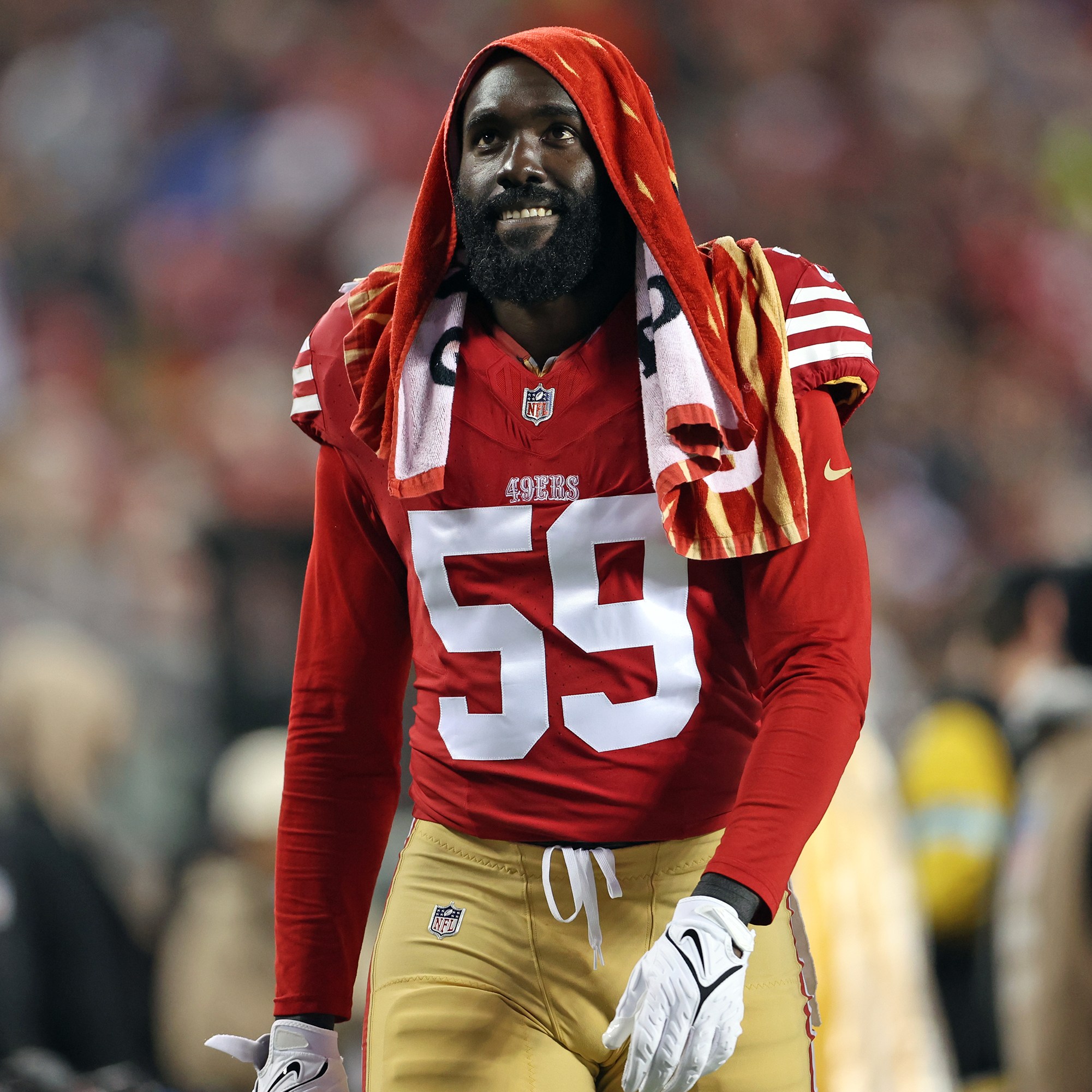 A football player wearing a red jersey with the number 59 stands on the sideline, a striped towel draped over his head. He is on a blurred field background.