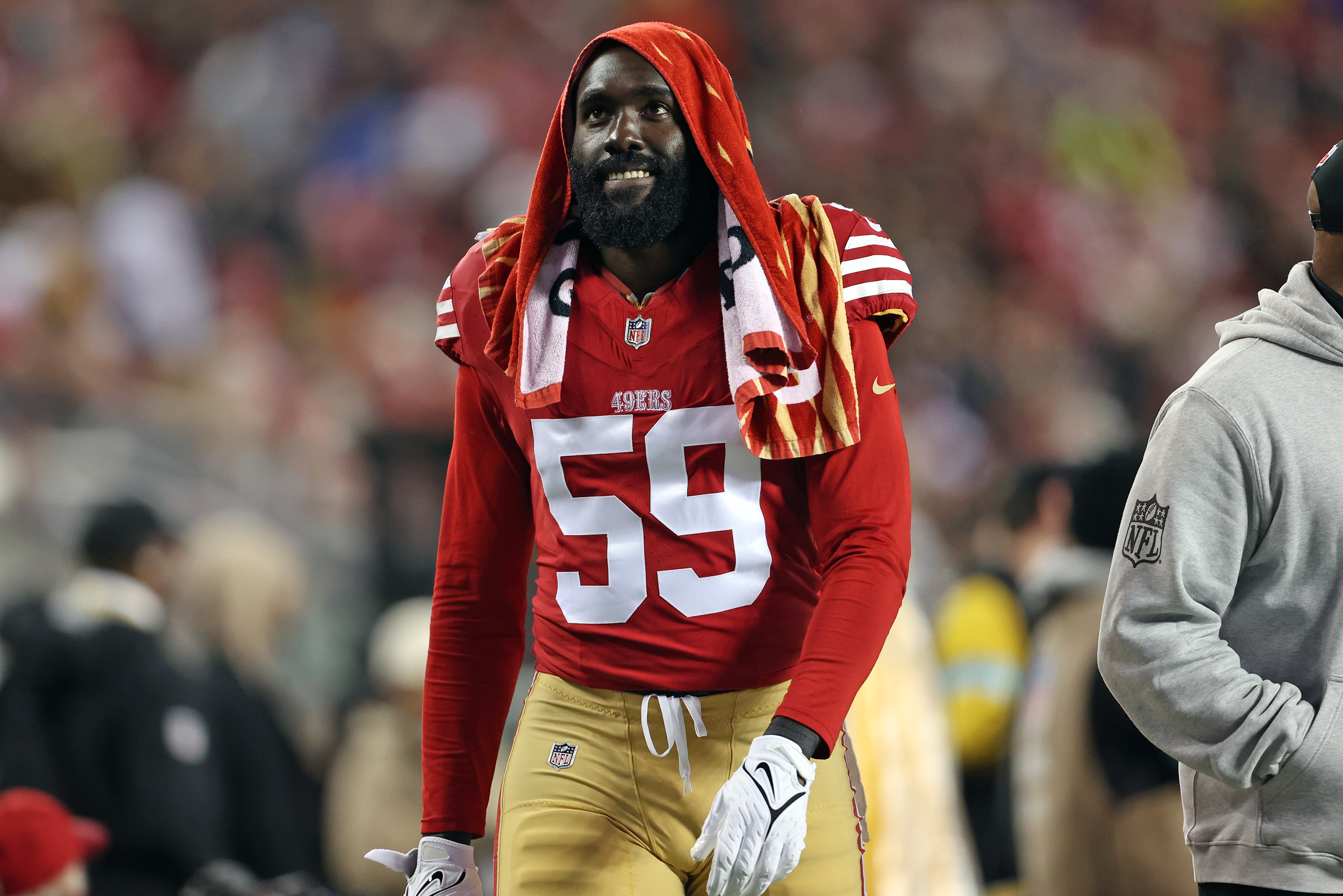 A football player wearing a red jersey with the number 59 stands on the sideline, a striped towel draped over his head. He is on a blurred field background.