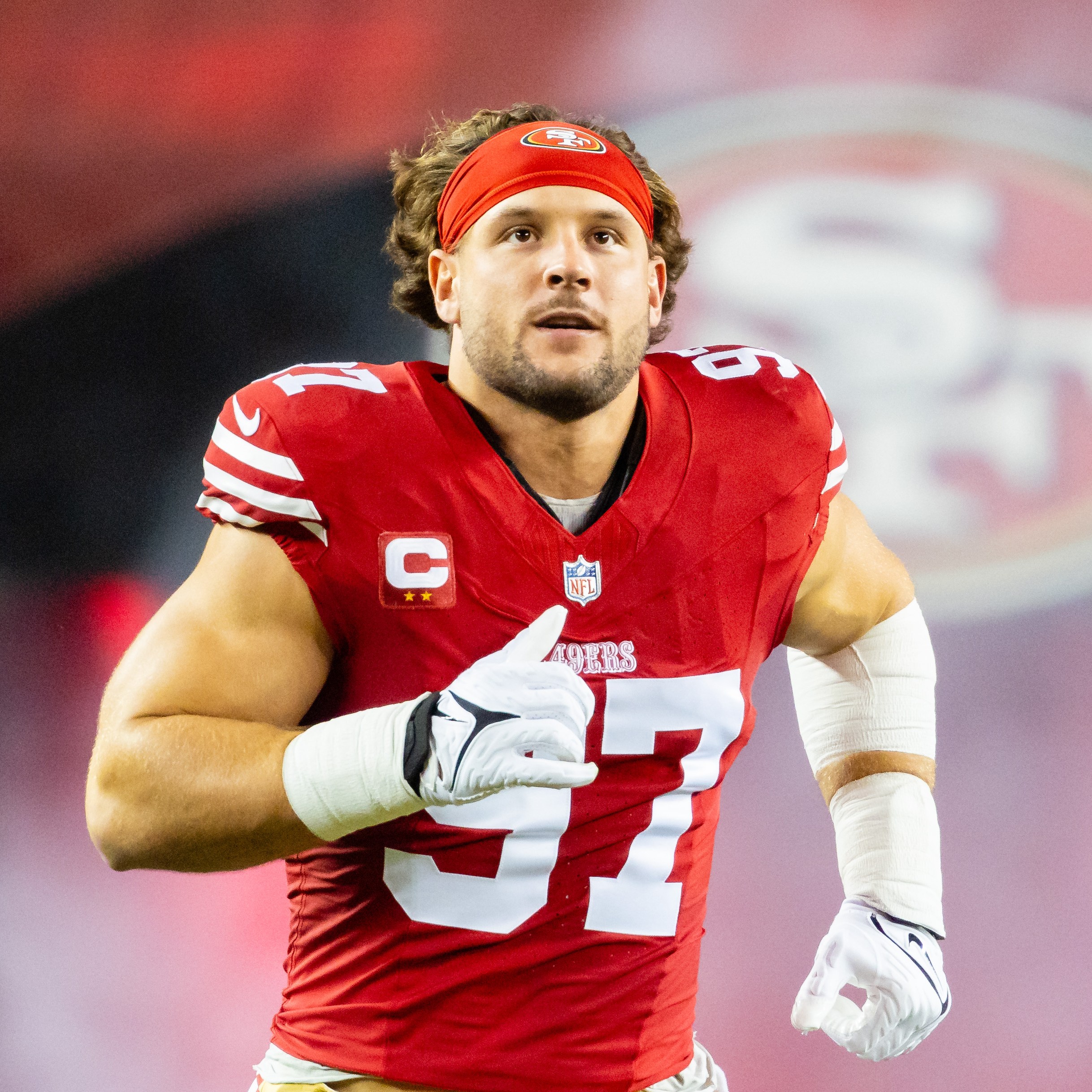 A football player in a red jersey with the number 97 is running on the field. He wears a red headband and has white gloves, with a large team logo in the background.