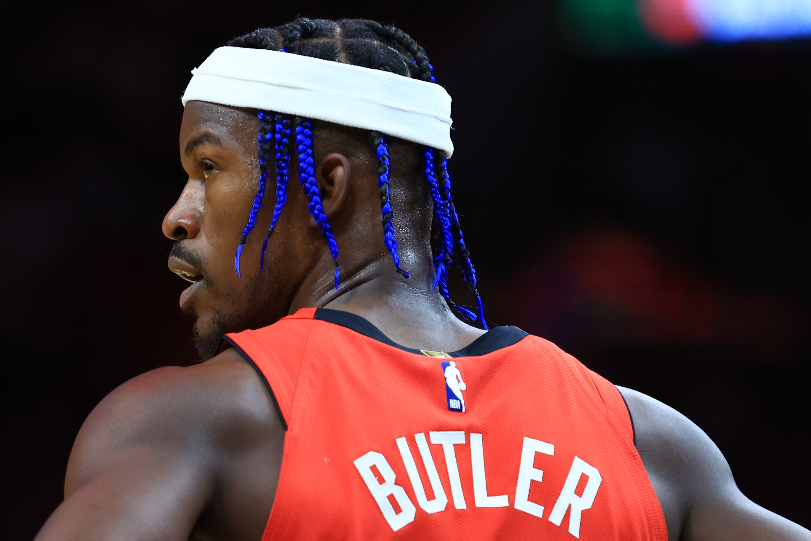 A basketball player with blue braided hair and a white headband is wearing a red jersey with &quot;BUTLER&quot; on the back. The NBA logo is visible on the jersey.