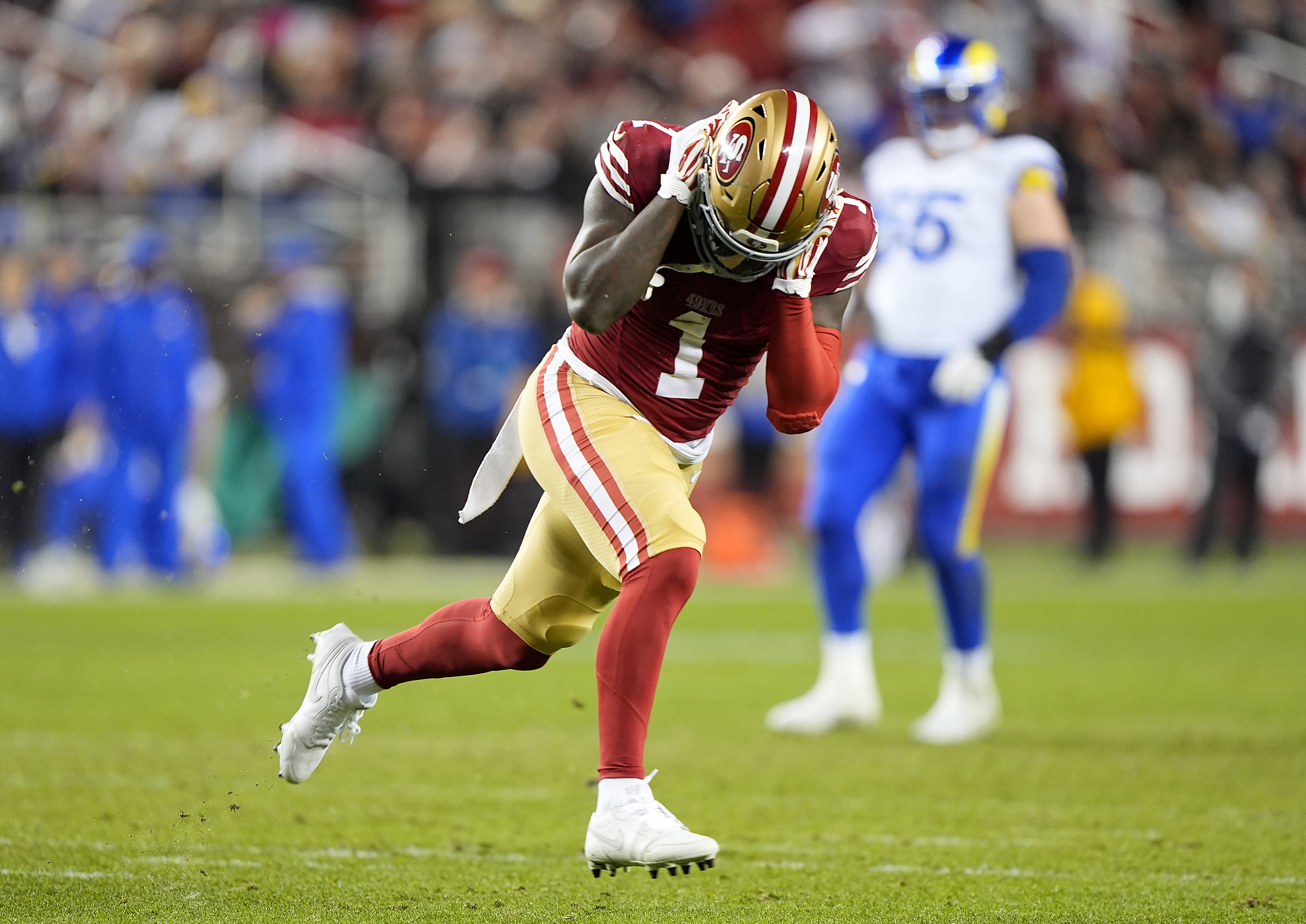 A football player in a red and gold uniform sprints with the ball, while another player in a white and blue uniform stands in the blurred background on the field.