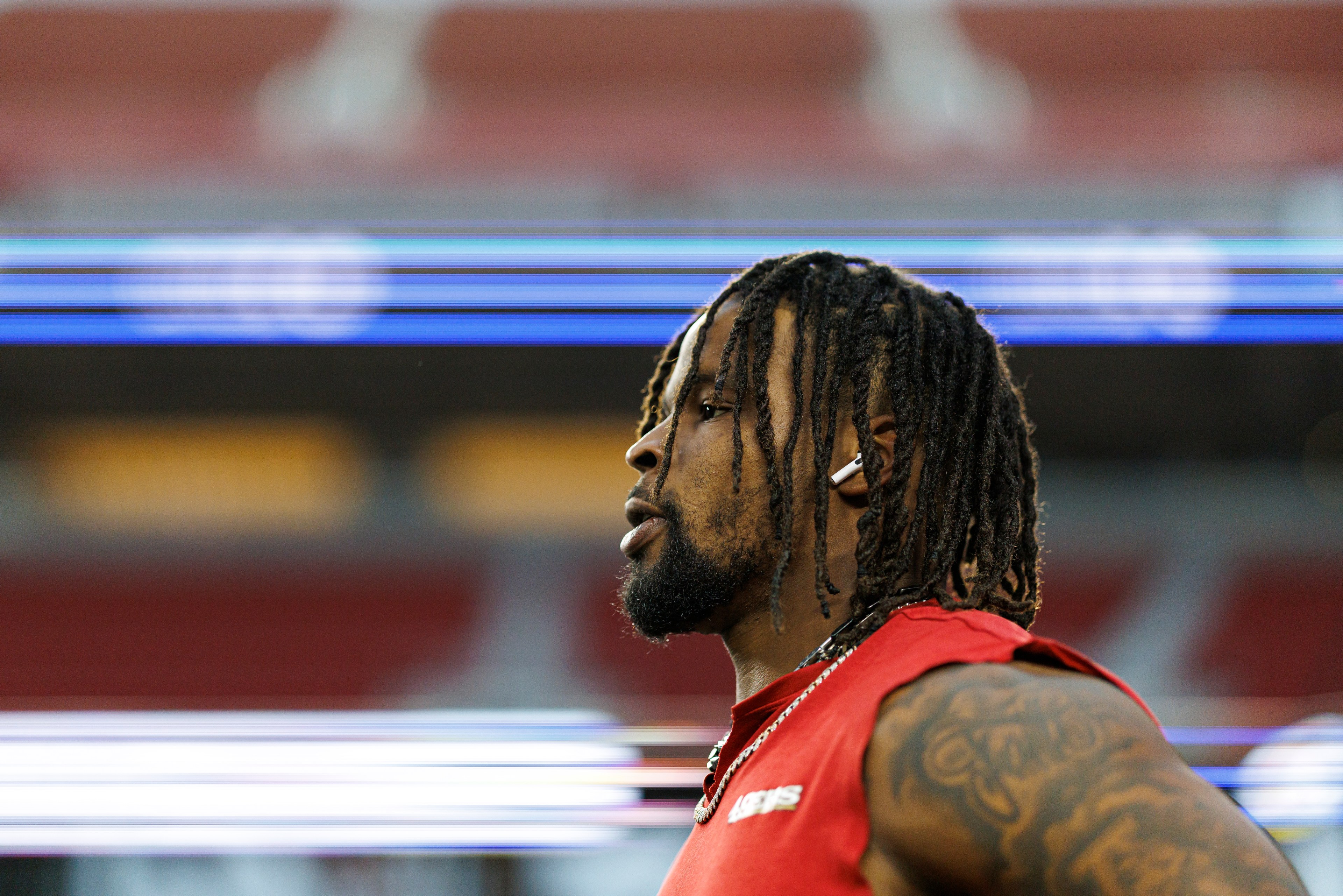 A person in profile with dreadlocks wearing a sleeveless red shirt and an earpiece stands against a blurred backdrop of lights and rows of seats.