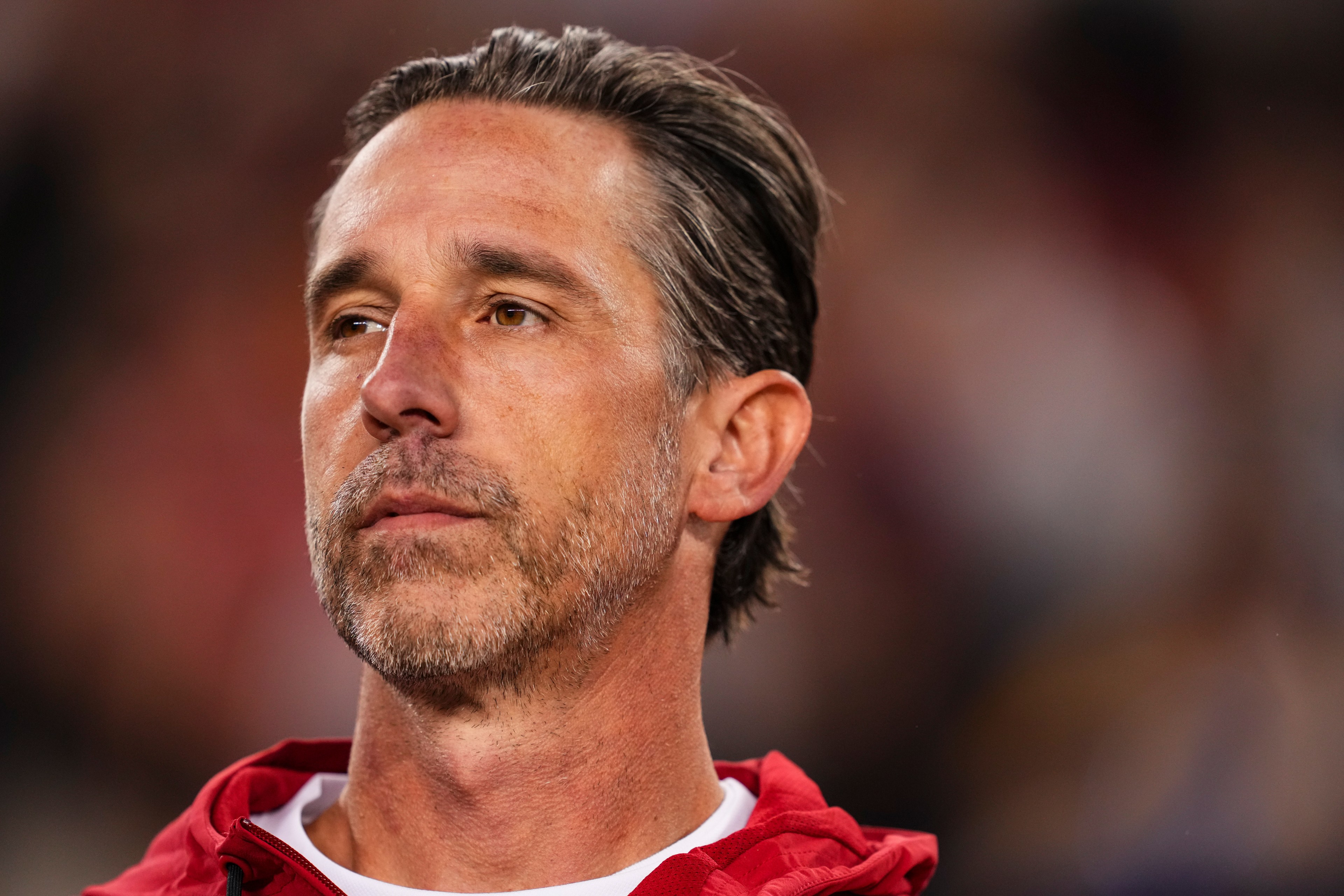 A man with short hair and a beard looks pensive, wearing a red jacket over a white shirt. The background is blurred, highlighting his thoughtful expression.