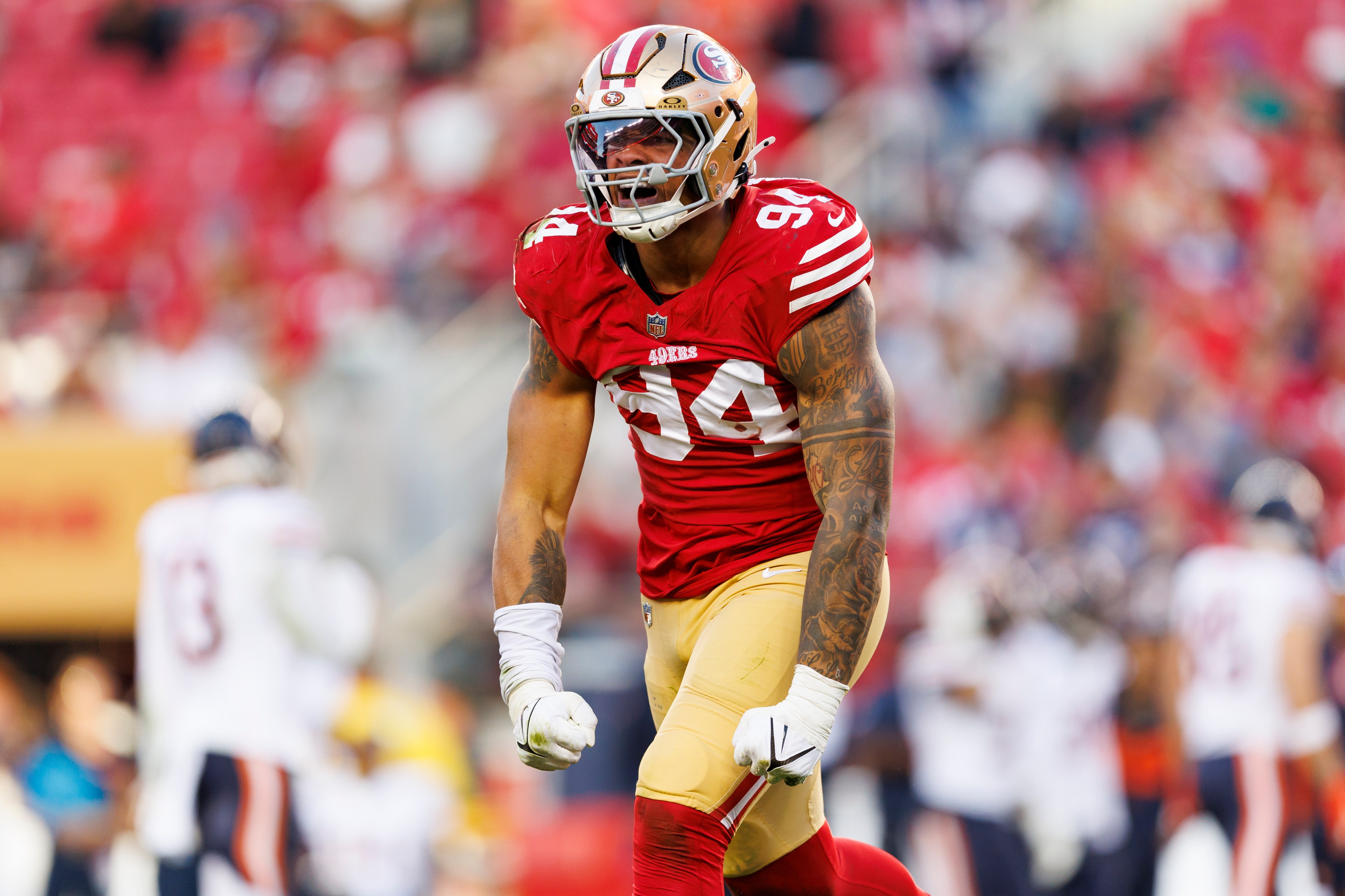 A football player in a red jersey with number 94, wearing a helmet and gloves, is clenching his fists on a field with blurred players in the background.