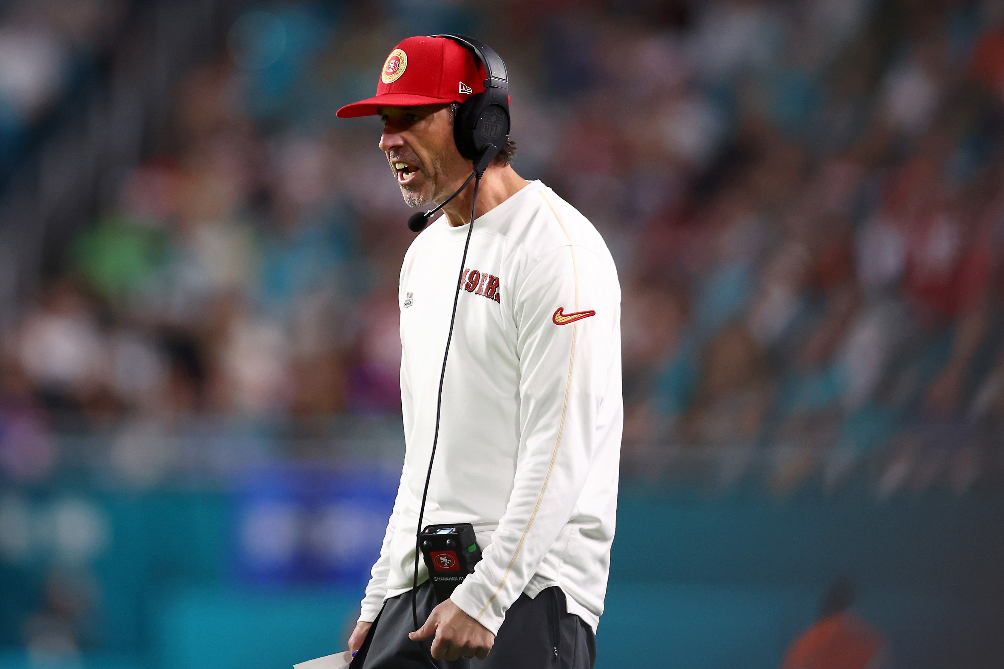 A man on a football field wears a red cap and headphones. Dressed in a white long-sleeve shirt with "49ers" on it, he appears to be shouting instructions.