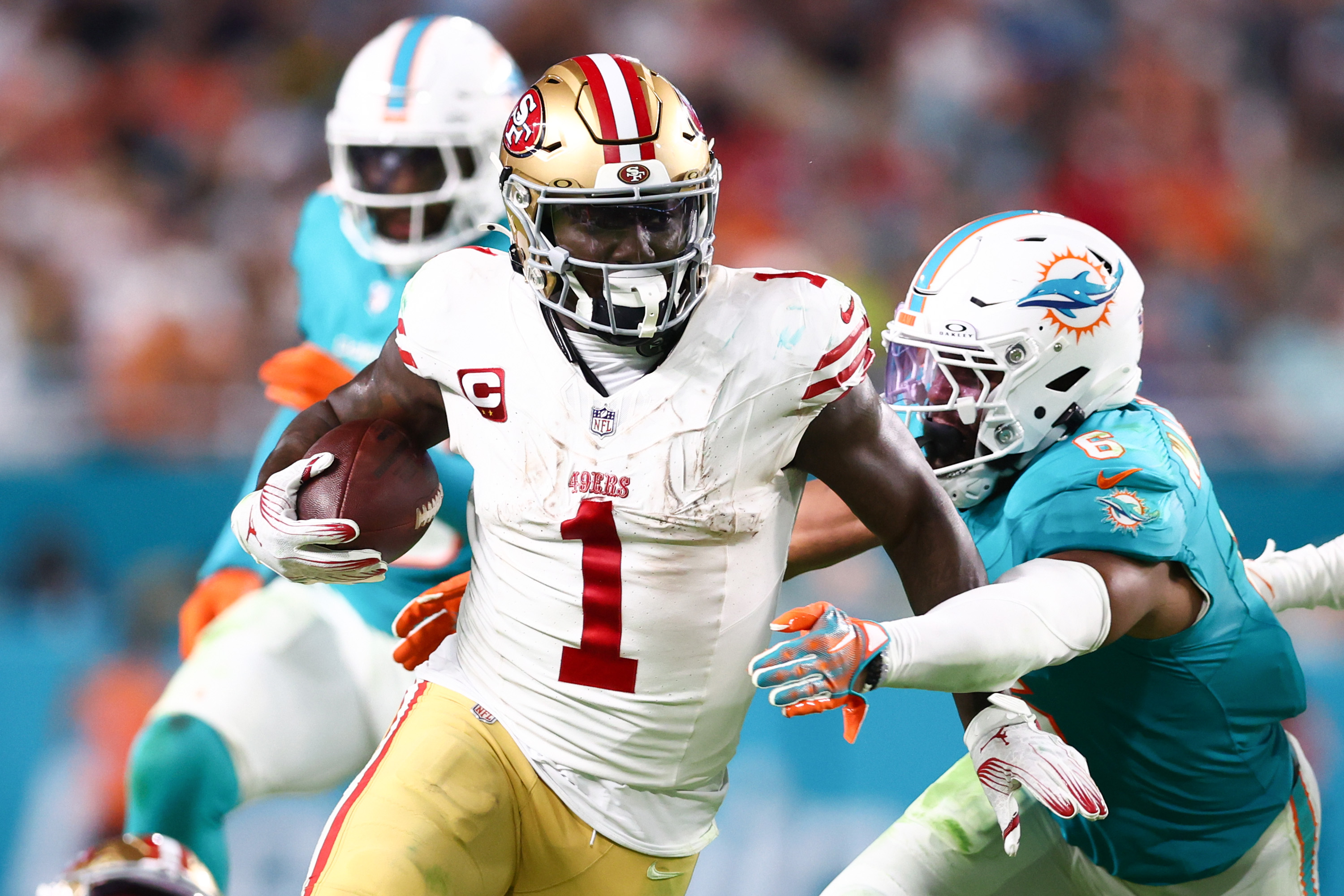 A football player in a white jersey runs with the ball while being pursued by players in teal jerseys on a field during a game.