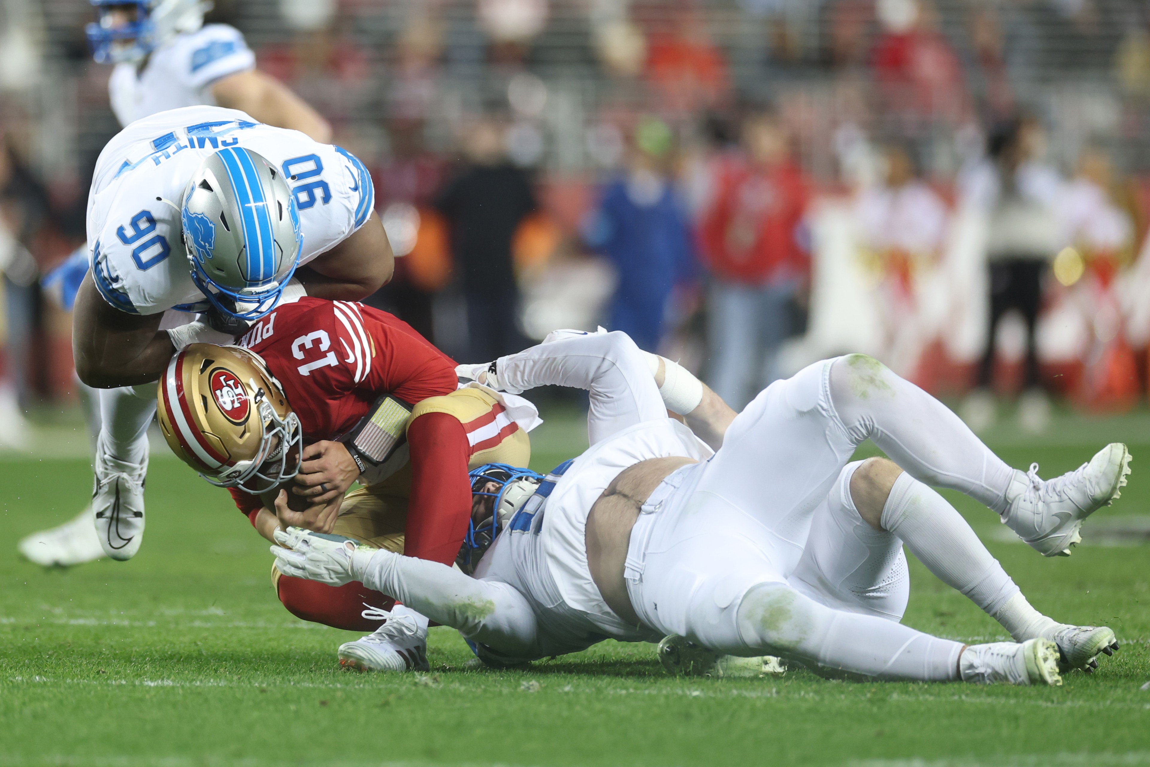 A football player in a red uniform is being tackled and brought to the ground by two players in white uniforms during a game, with a focused crowd in the background.