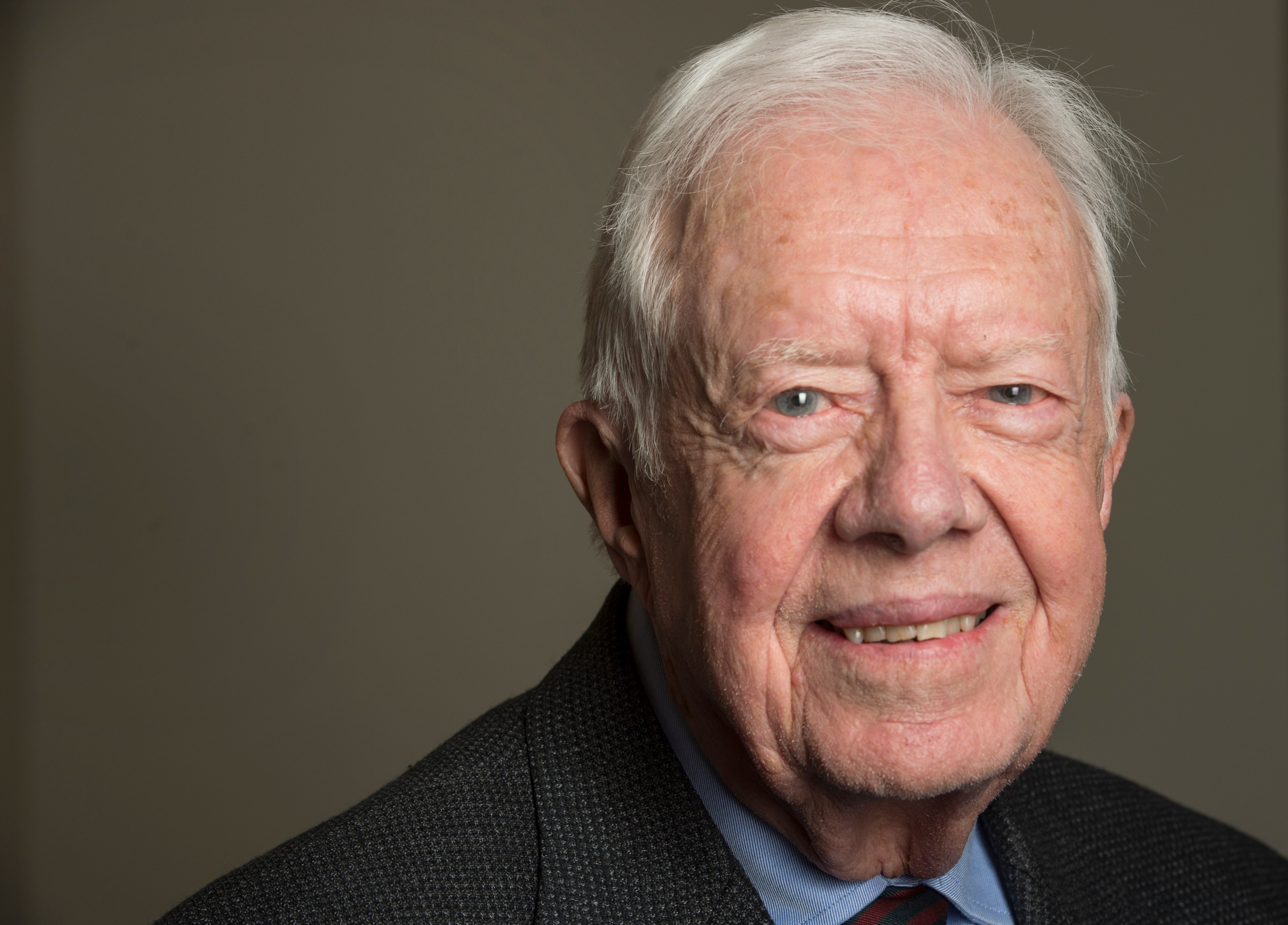 An elderly man with white hair is smiling slightly. He is wearing a dark jacket, light blue shirt, and a patterned tie against a plain background.