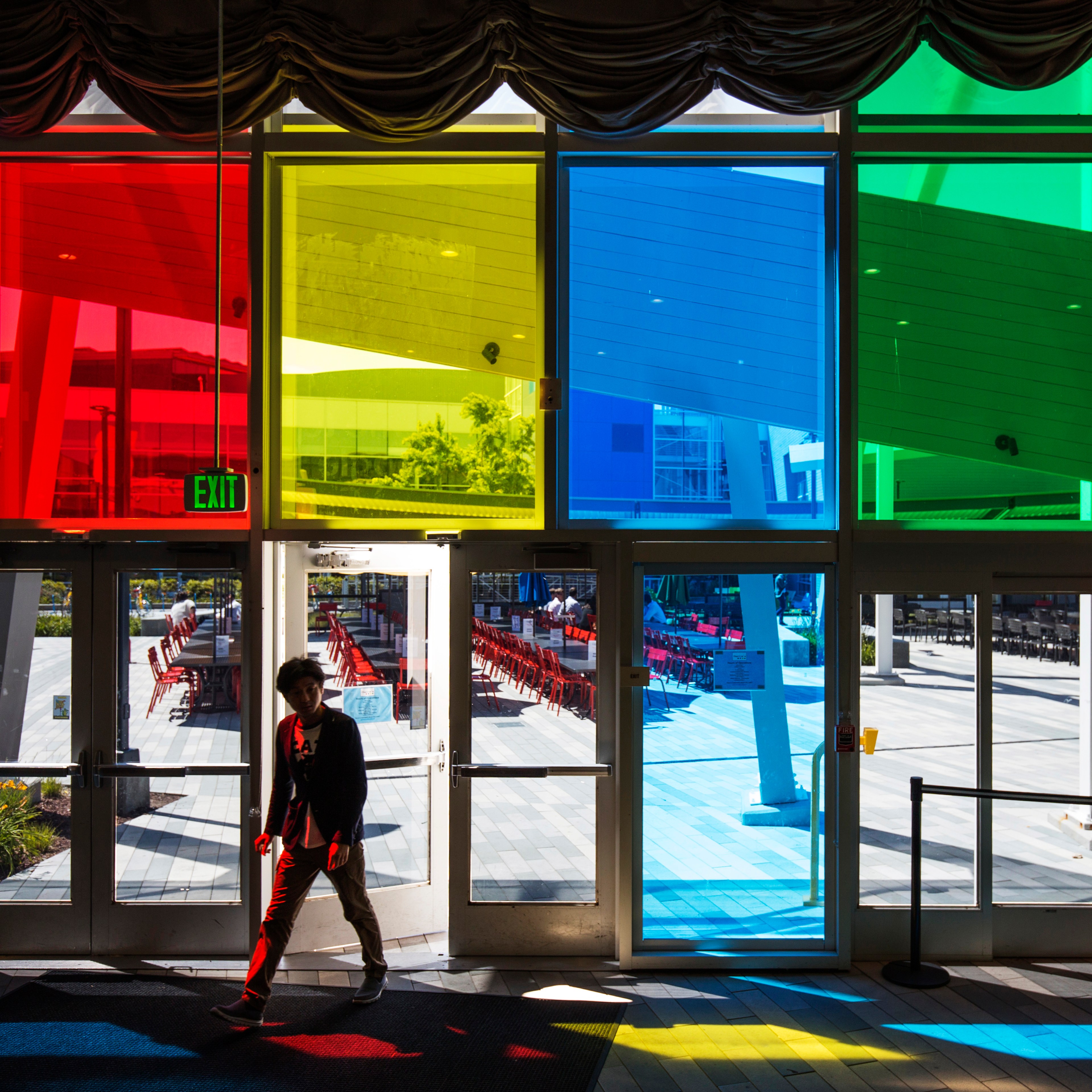 A person walks inside through glass doors with colorful window panels casting red, yellow, blue, and green hues on the ground. An "Exit" sign hangs above.