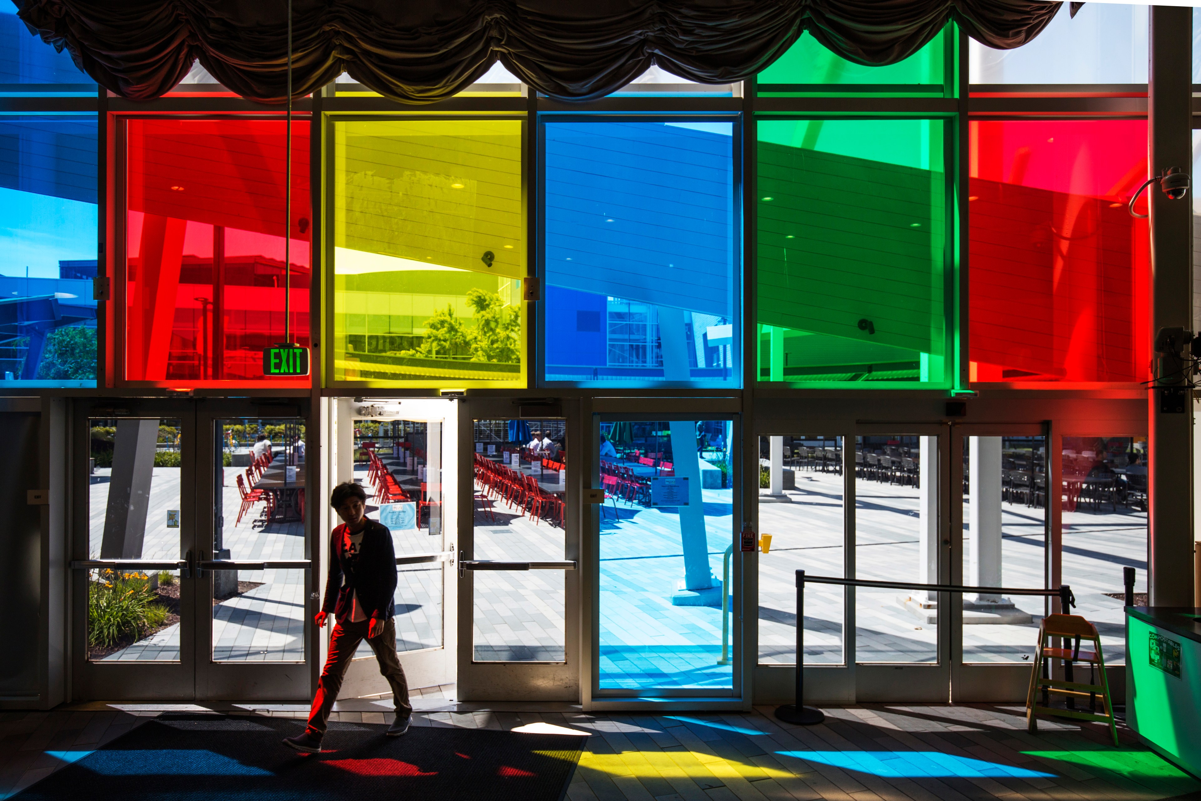 A person walks inside through glass doors with colorful window panels casting red, yellow, blue, and green hues on the ground. An "Exit" sign hangs above.